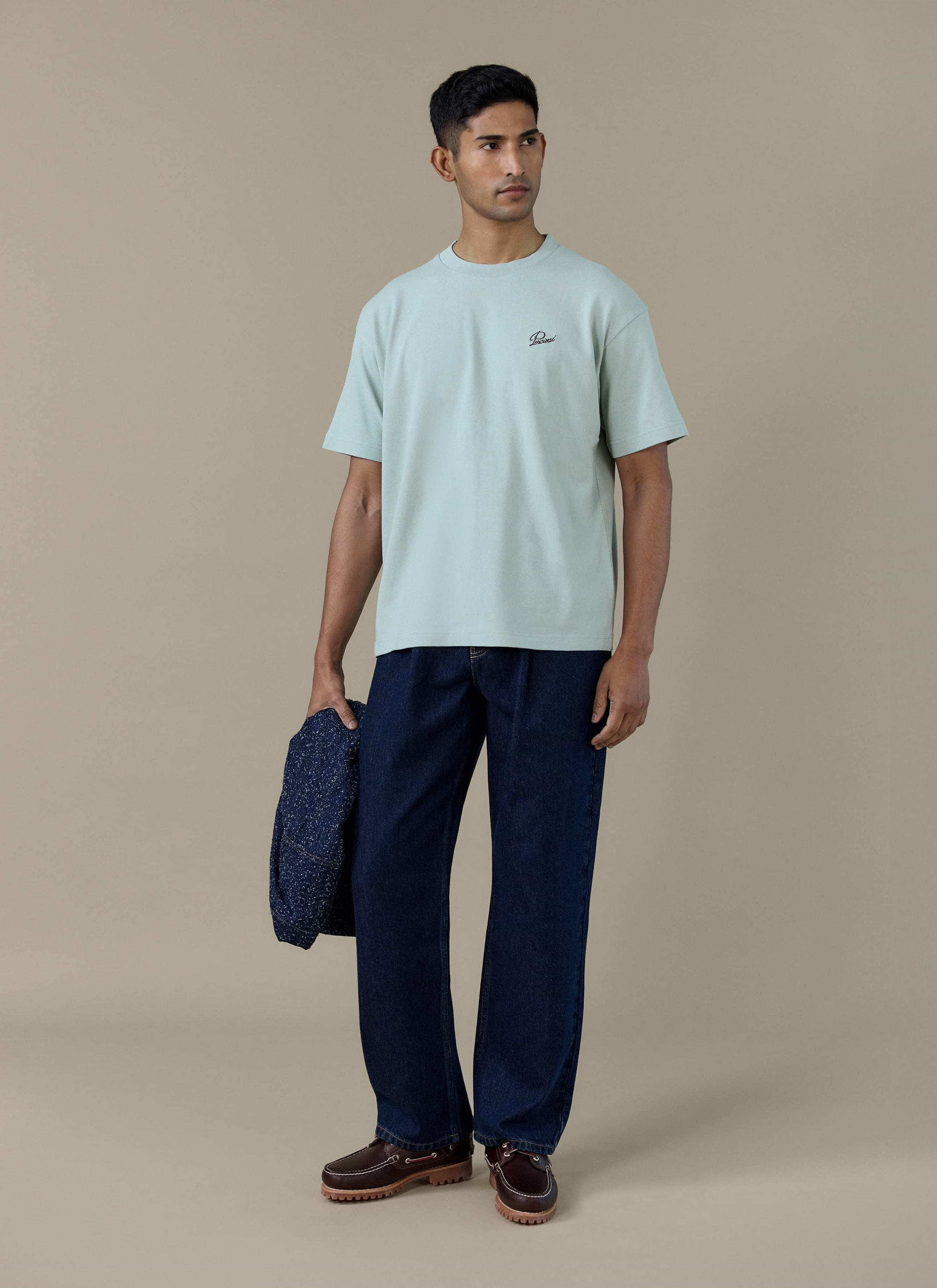 A model wearing the sage green Percival Perci Script T Shirt in organic cotton, styled with navy trousers.