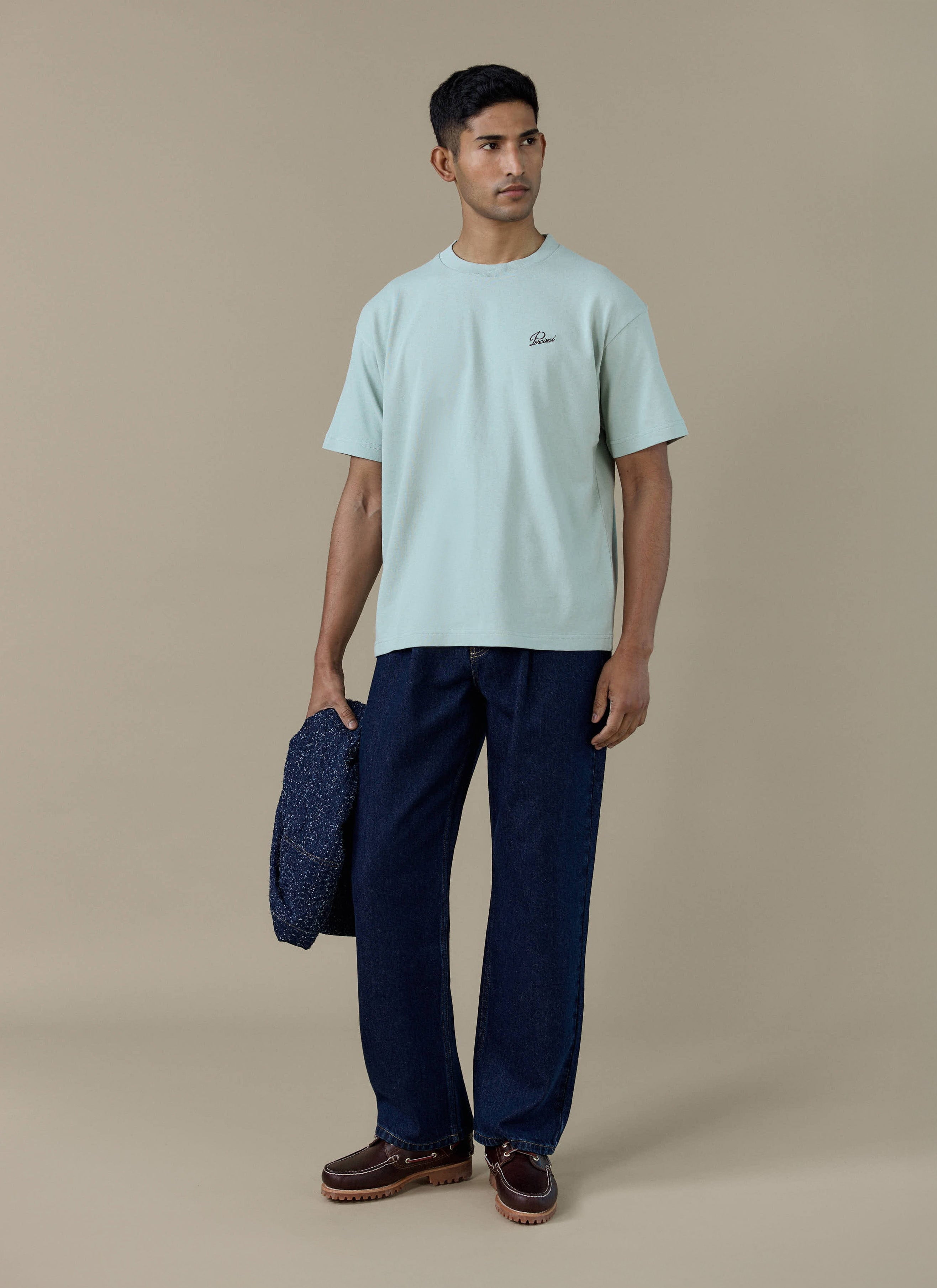 A model wearing the sage green Percival Perci Script T Shirt in organic cotton, styled with navy trousers.
