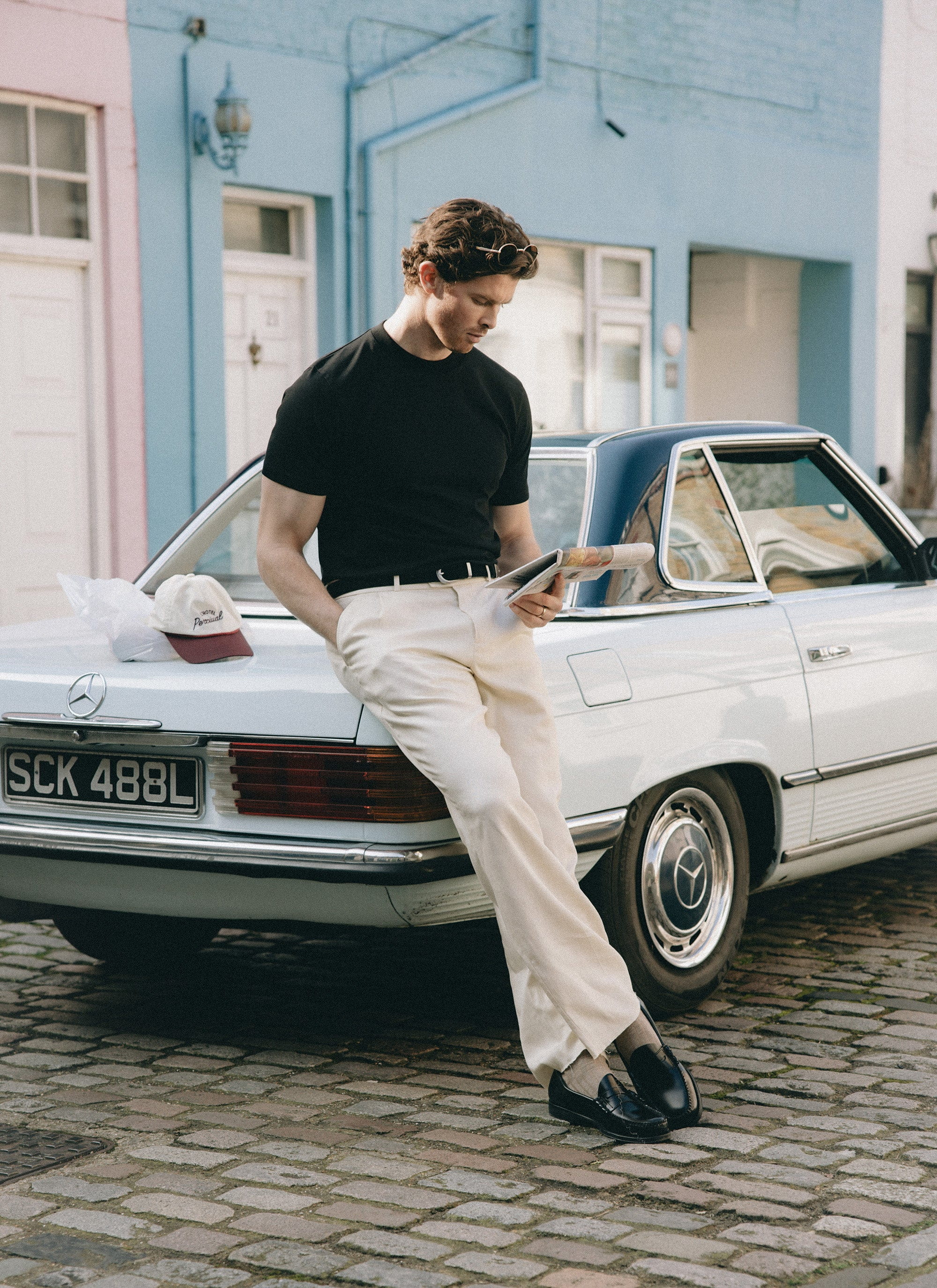A model wearing a plain black Percival t-shirt and white trousers, leaning against a vintage car reading a newspaper.