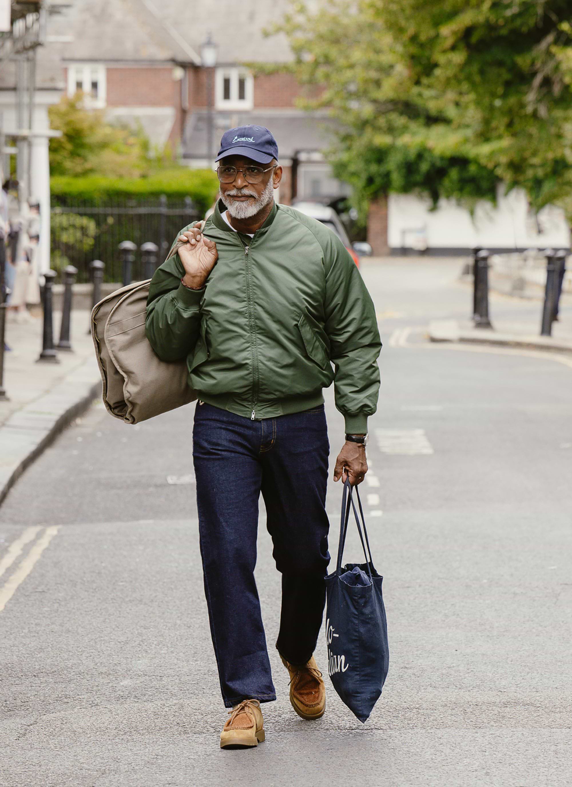 A man models the Percival Nylon Bomber Jacket in Forest Green, walking down a street carrying a tote and garment bag.