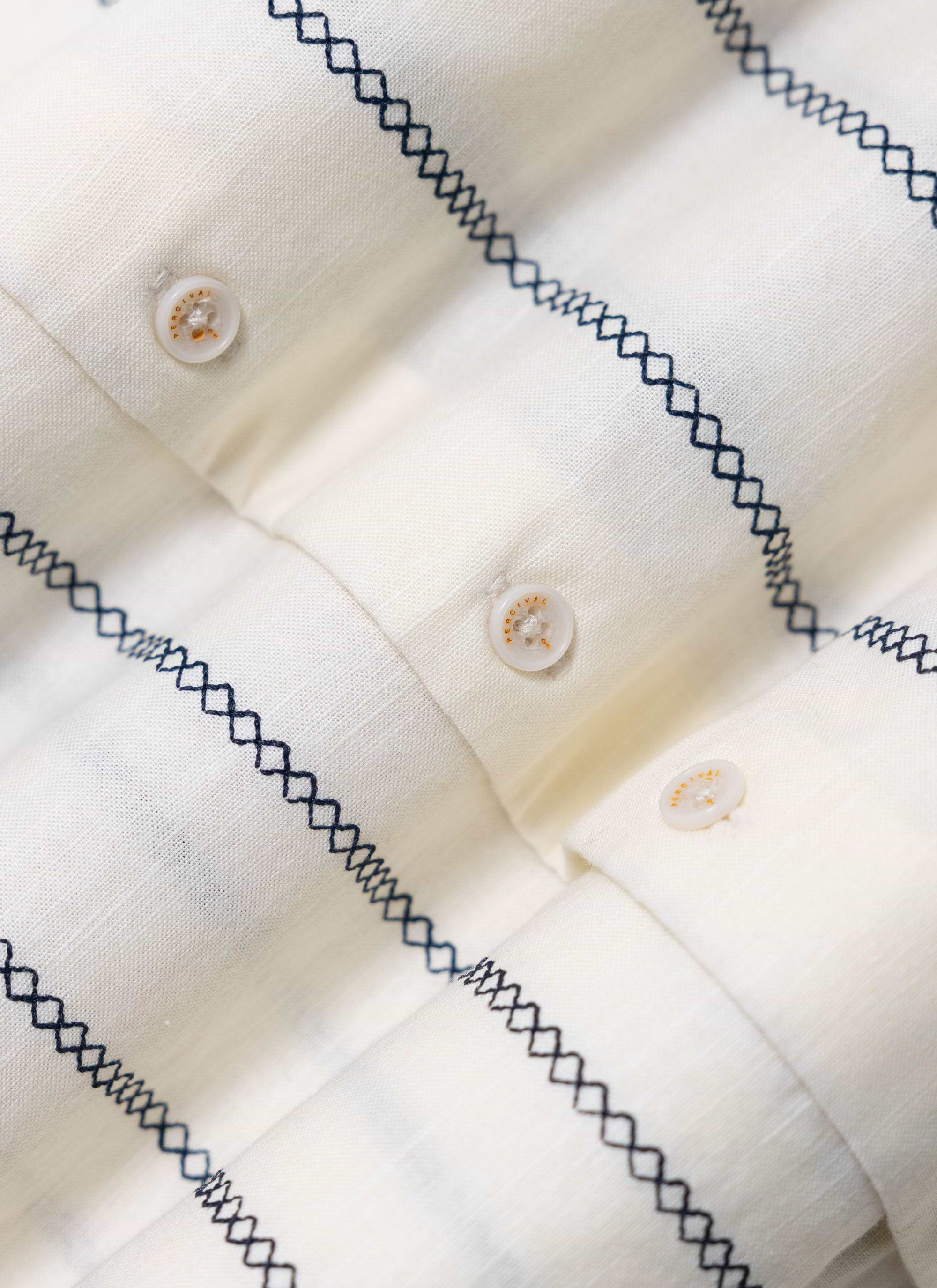A close-up of the Percival Cross Stitch Boxy Clerk Shirt in ecru, detailing the blue stitch pattern and custom buttons.