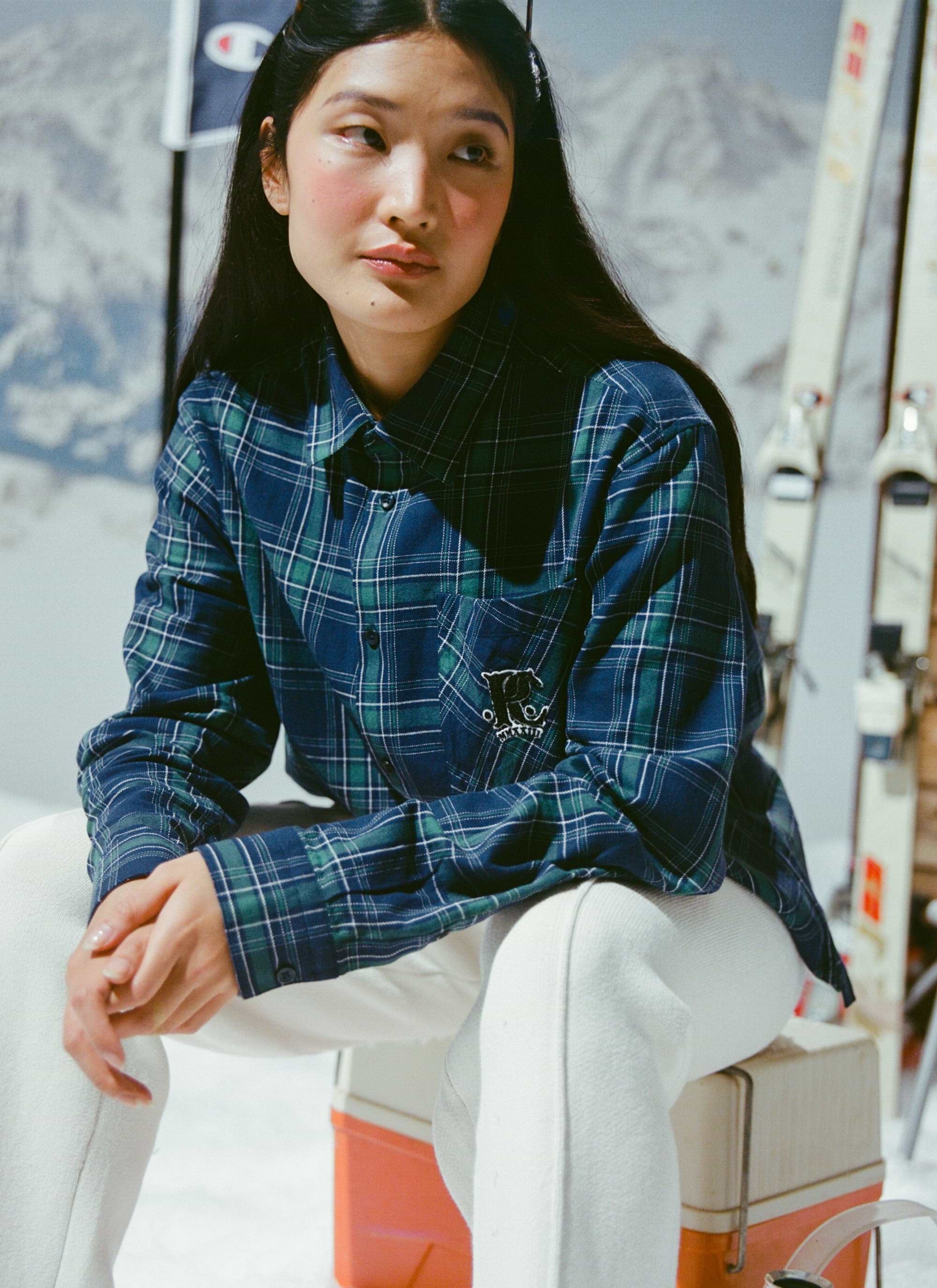 A woman models the Percival x Champion Check Boxy Long Sleeve Shirt in a blue and green tartan against a ski backdrop.