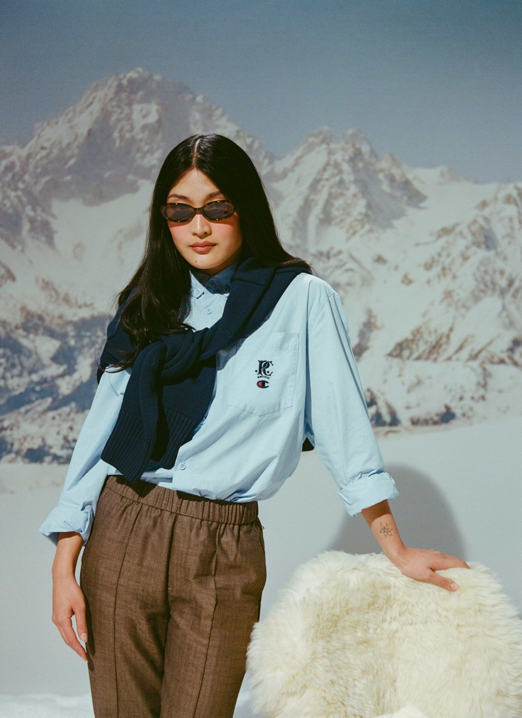 A model wears the light blue Champion x Percival Relaxed Oxford Shirt with a navy jumper draped over her shoulders.
