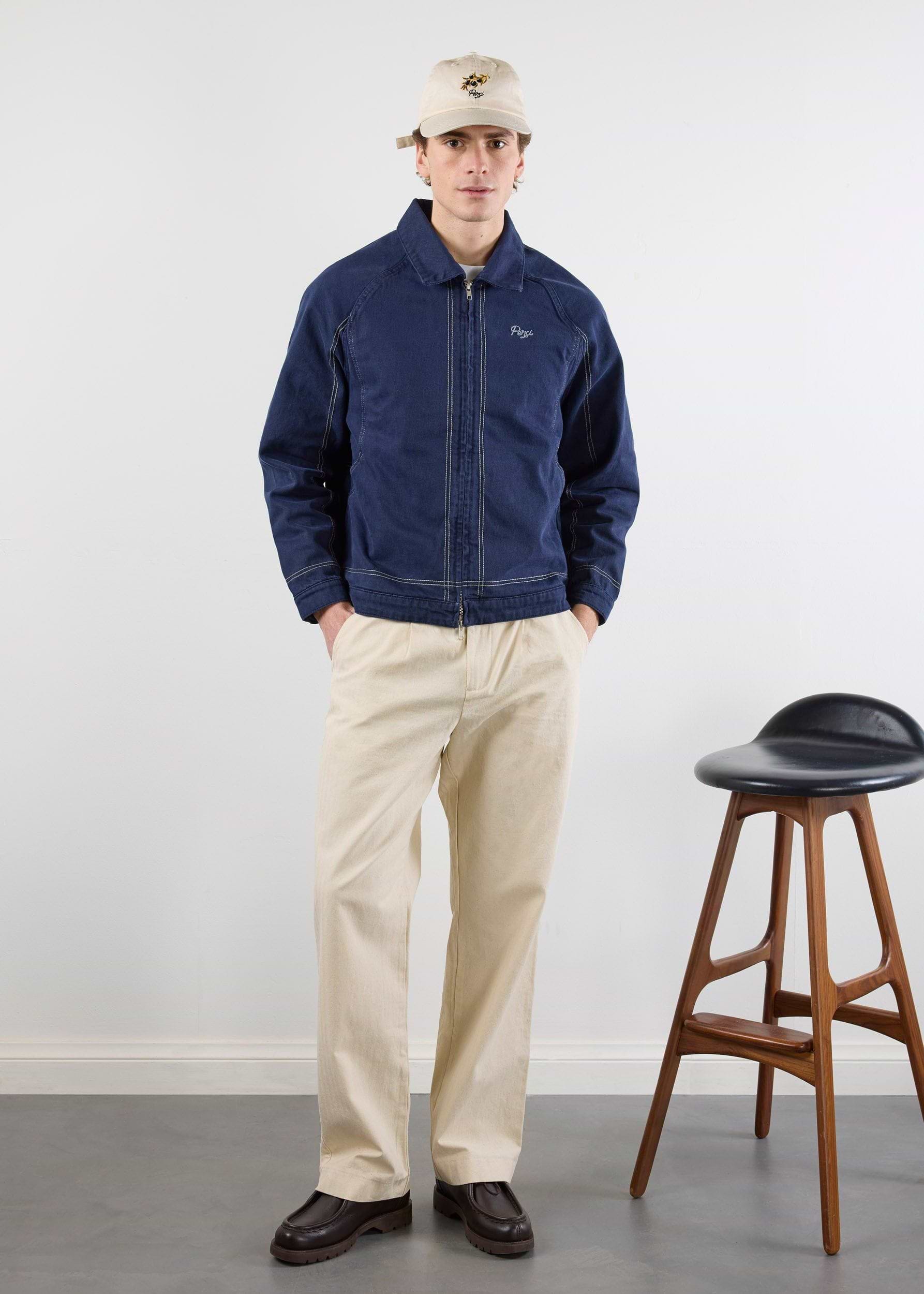 A model wearing the navy Percival Calabria Harrington Jacket with contrast stitching, styled with cream trousers and a cap.