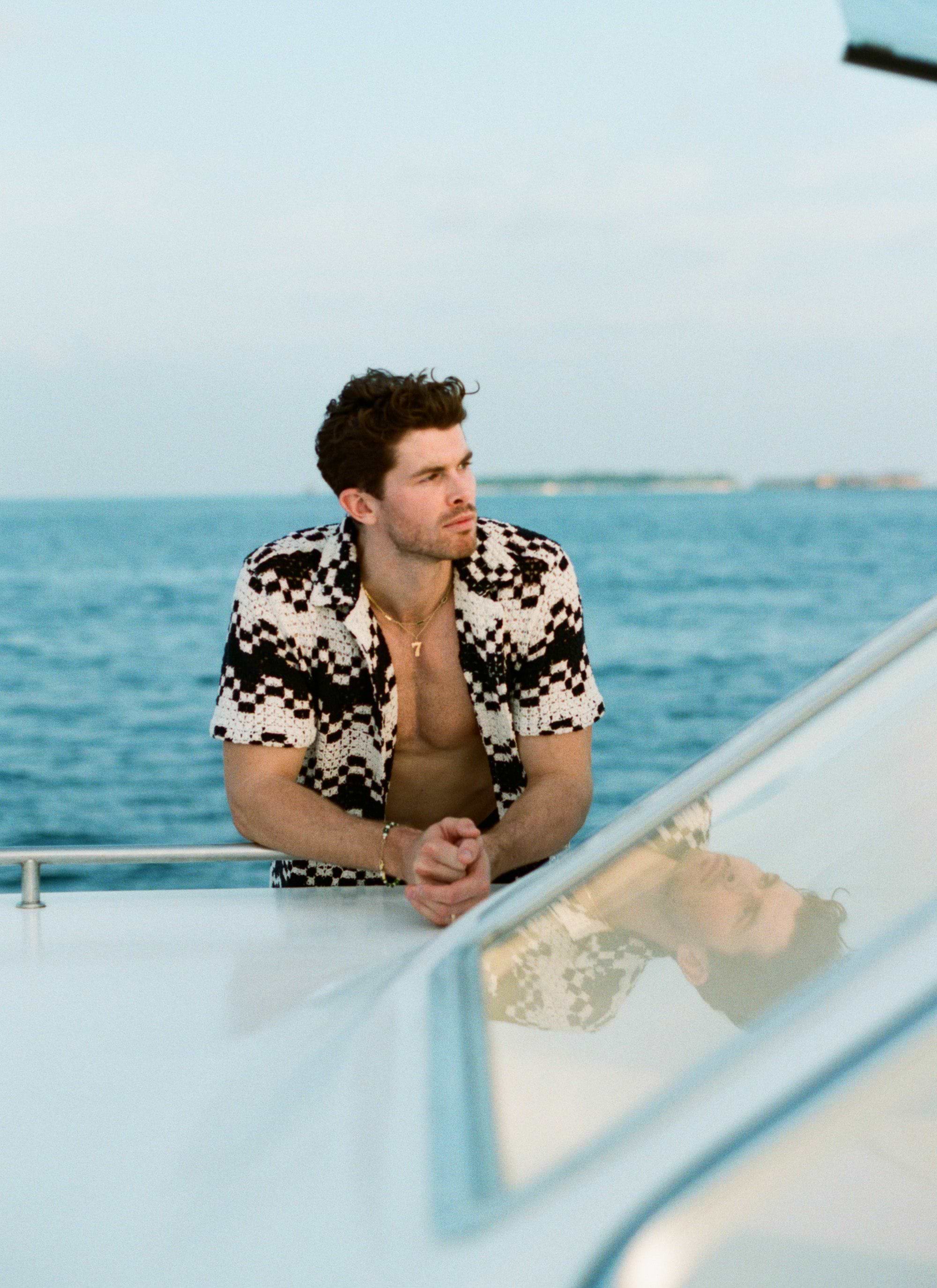 A man on a boat models the Percival Naples Crochet Cuban Shirt in a black and ecru geometric pattern against the ocean.