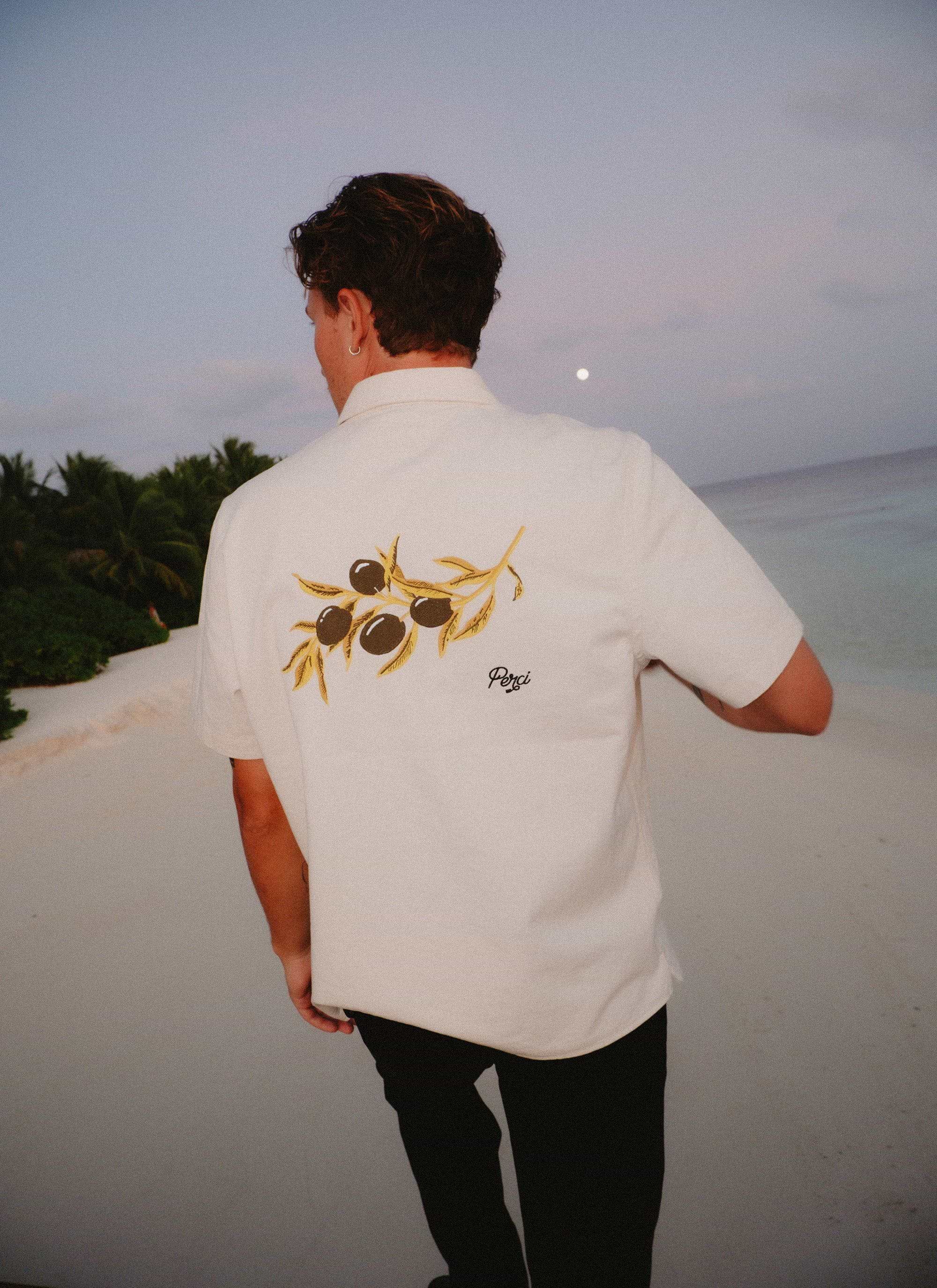 A man on a beach at dusk wearing the white Percival Olive Embroidered Cropped Shirt, showing the olive branch embroidery.