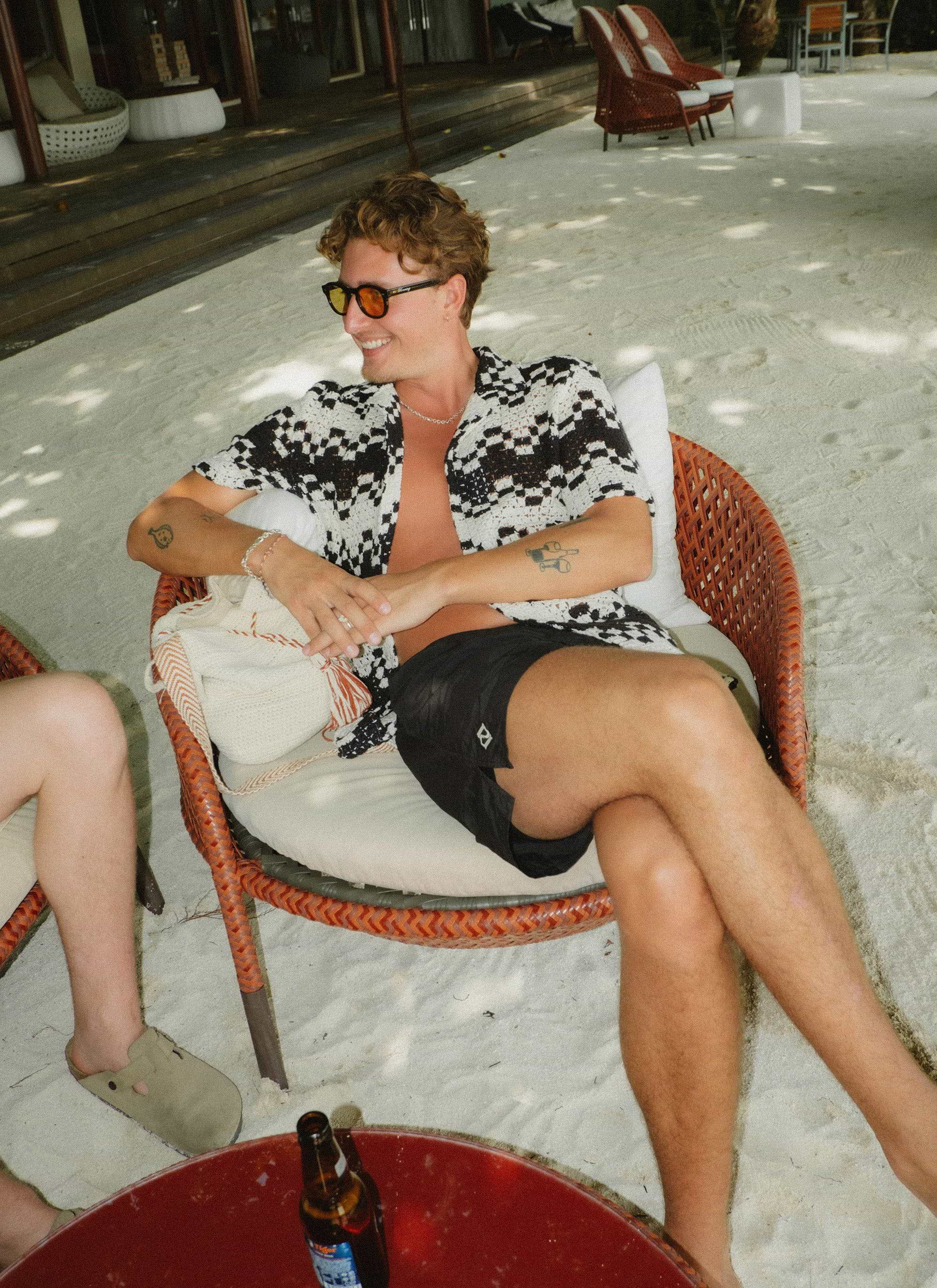 A man relaxing on a beach chair models the black and ecru patterned Naples Crochet Cuban Shirt from Percival.