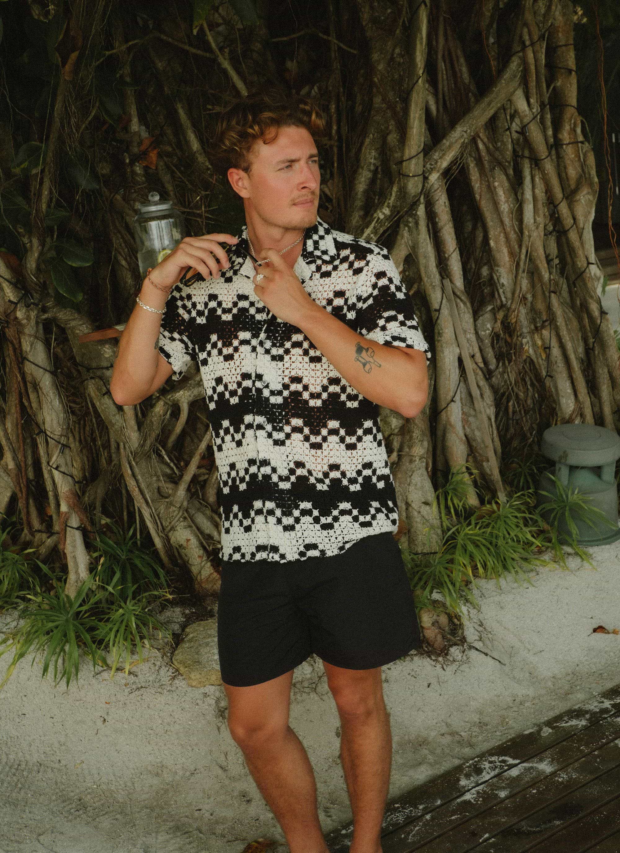 A man models the Percival Naples Crochet Cuban Shirt in ecru and black, adjusting the collar while posing by a large tree.