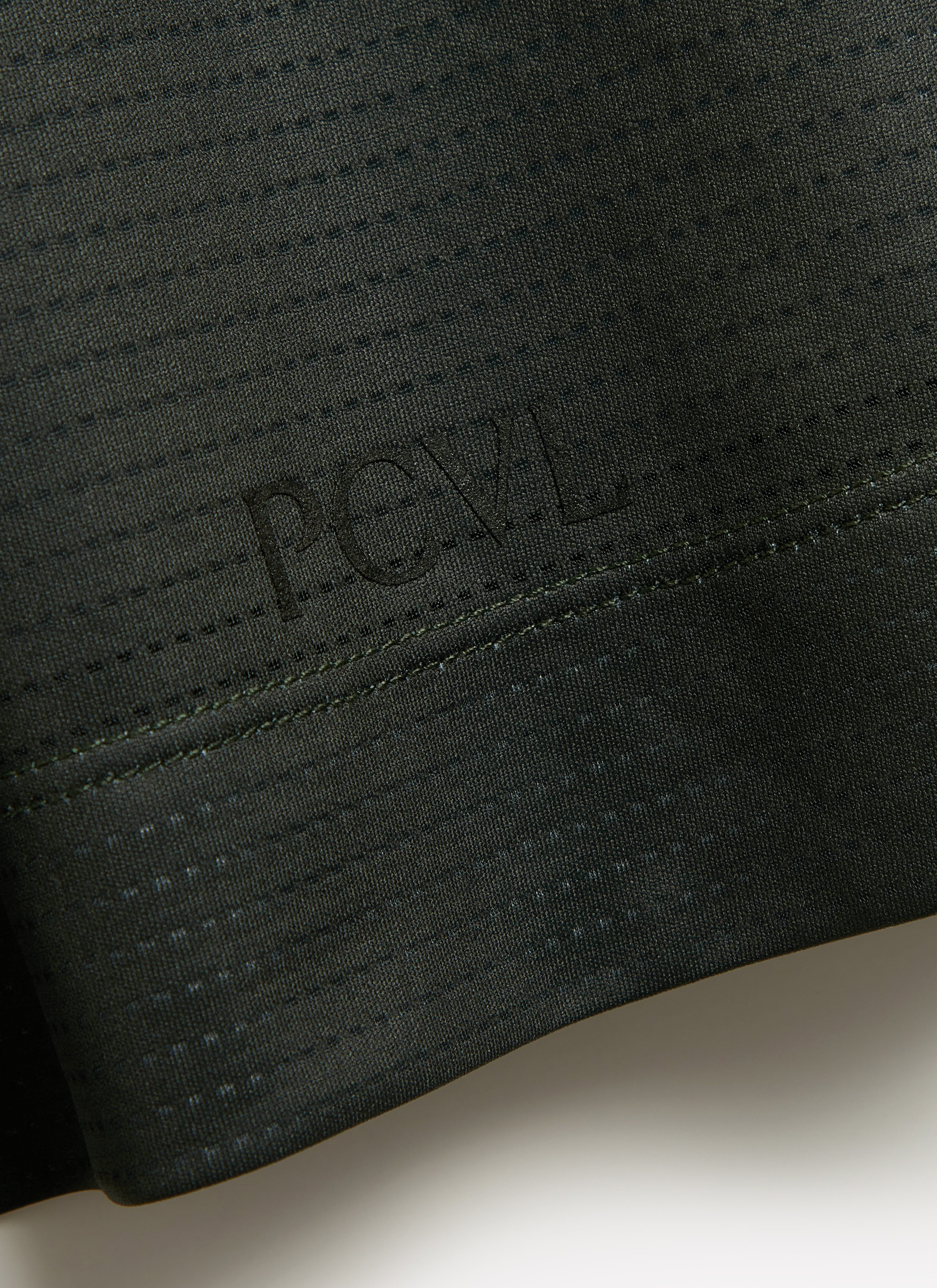 A close-up of the tonal PCVL logo embossed on the hem of the forest green Performance Top by Percival.