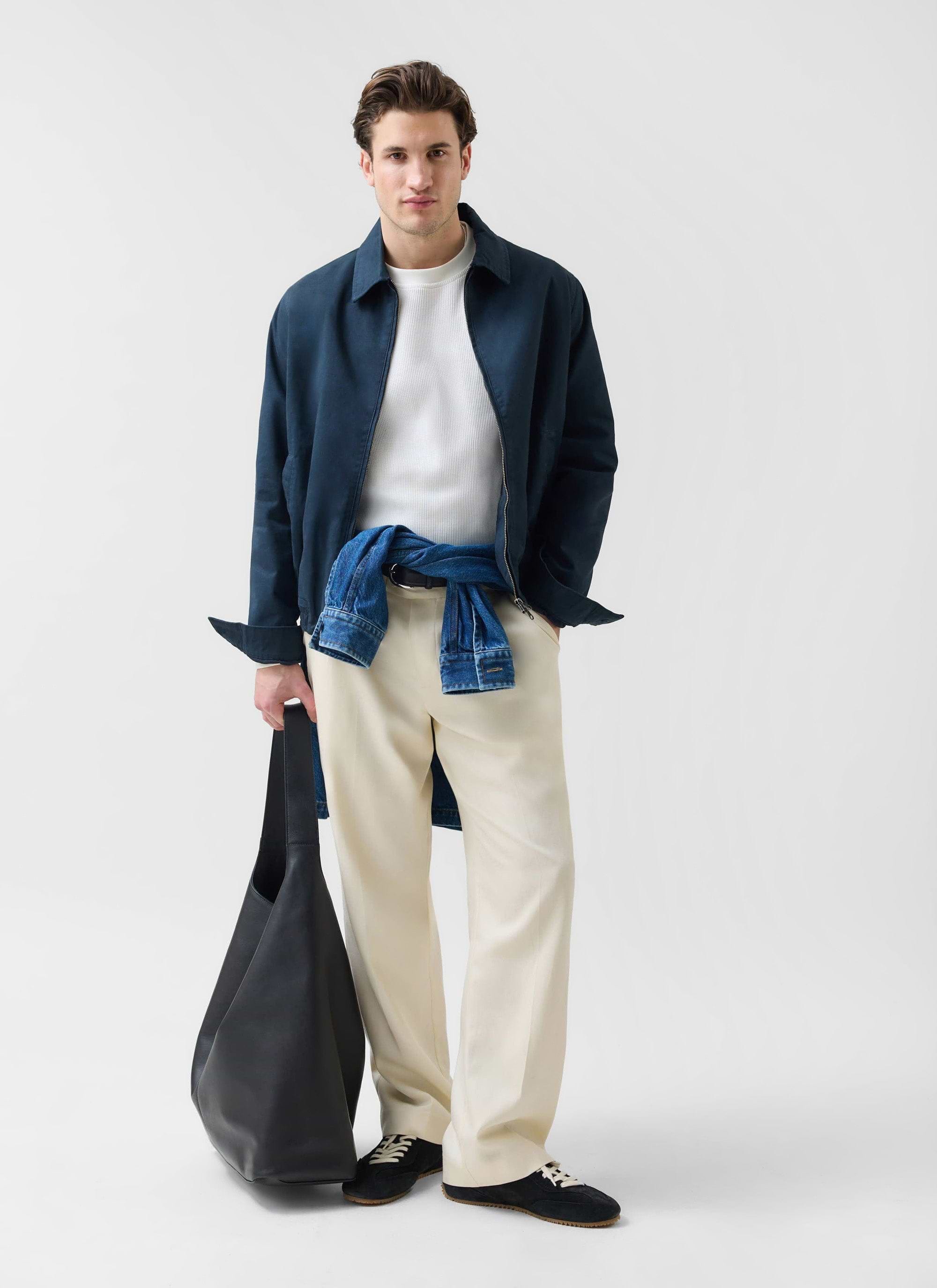 A model wears the navy Percival Leo Harrington jacket with cream trousers, a denim jacket tied at his waist, and a tote bag.