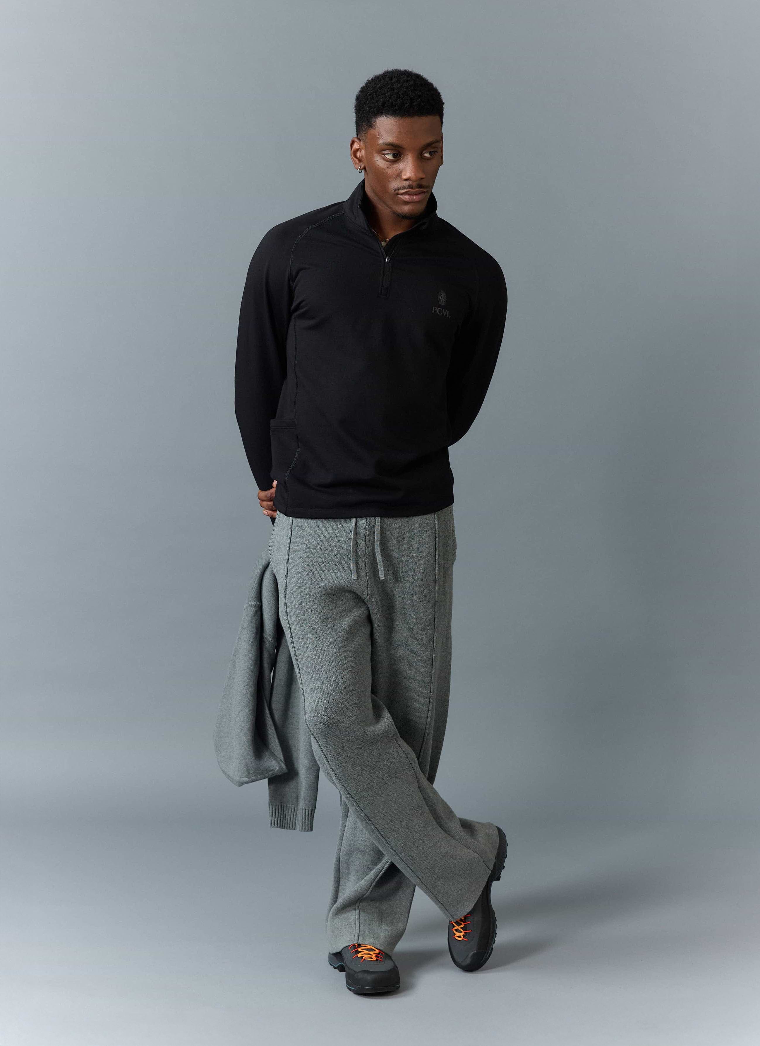 A model wears the black PCVL Base Layer Quarter Zip Jacket from Percival, styled with grey trousers and black walking shoes.