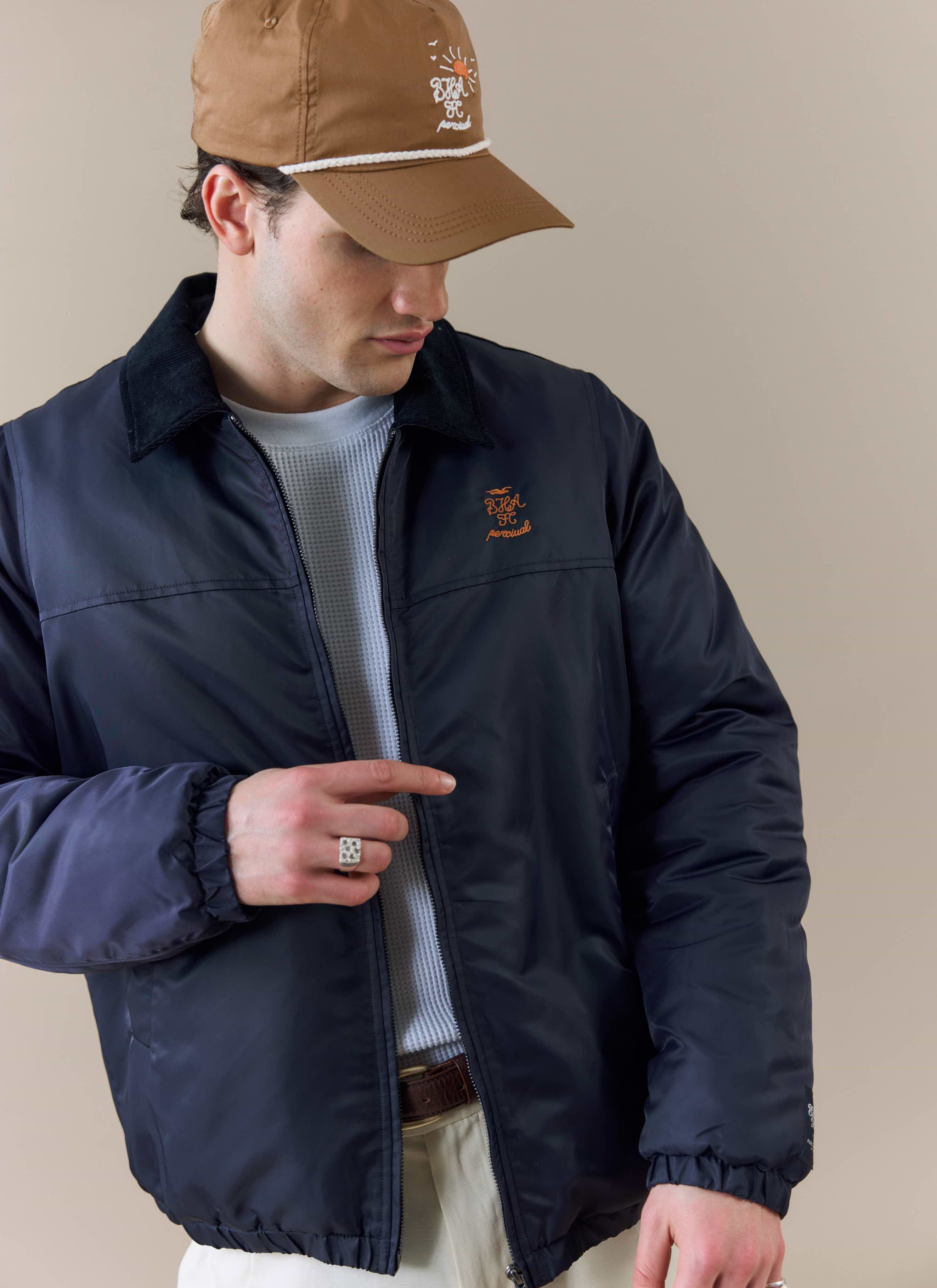 A model wears the Percival x Brighton & Hove Albion F.C. Coach Jacket in navy, showing the orange chest embroidery.