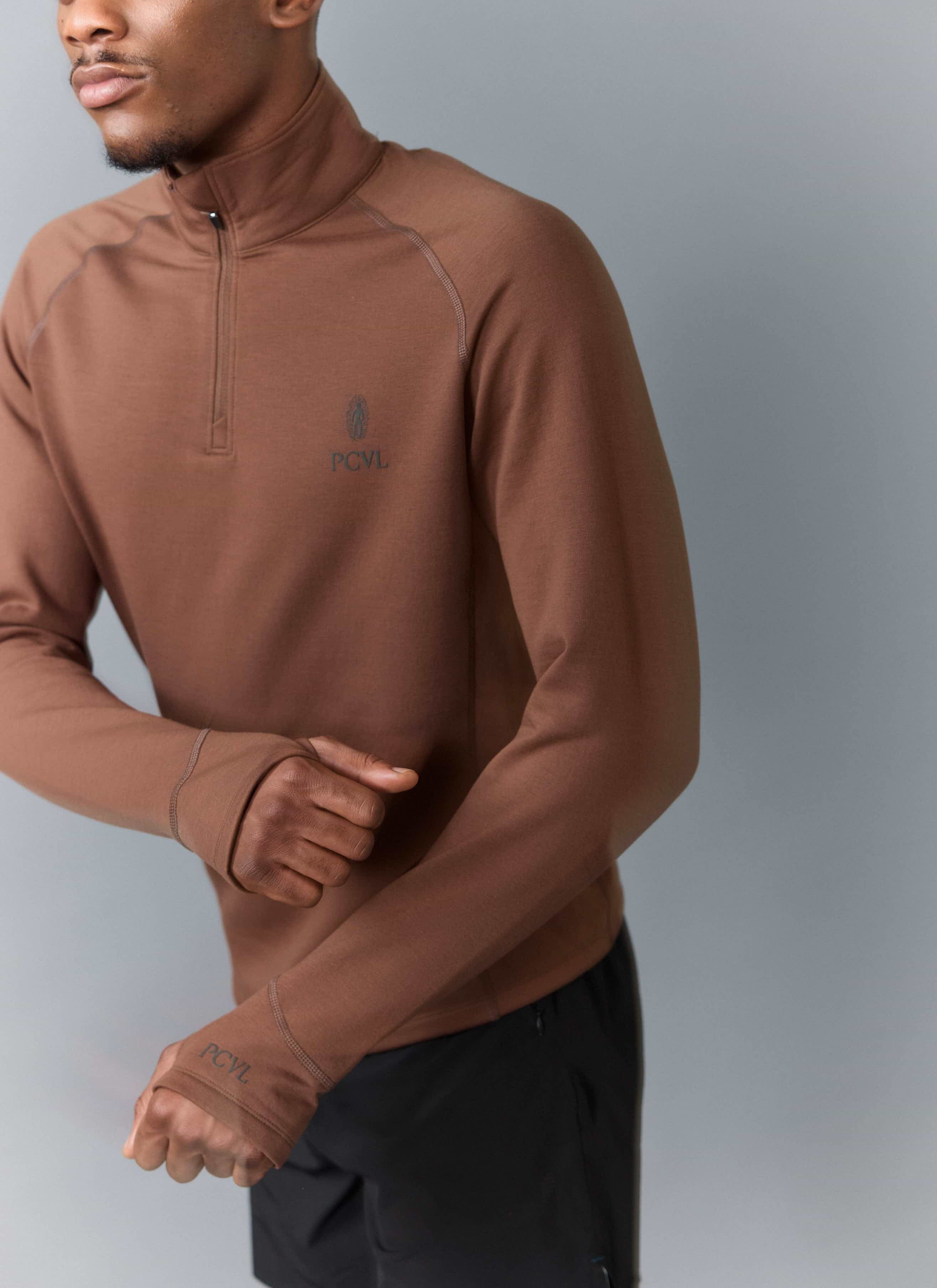 A close-up of a model wearing the Percival PCVL Base Layer Quarter Zip in rust, showing the chest logo and thumb loops.
