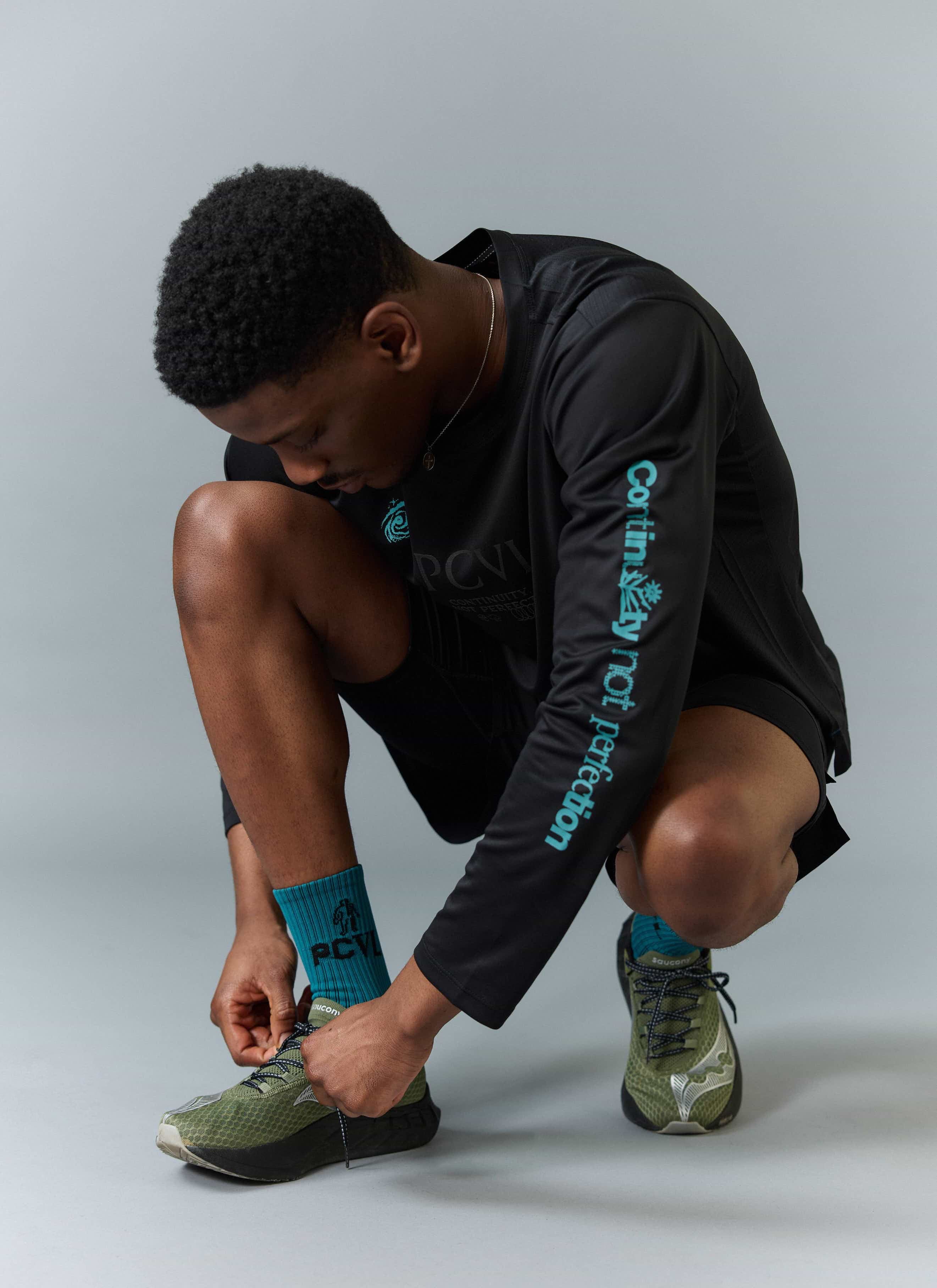 A model wearing the Percival black long sleeve PCVL performance top, crouching to tie the laces on his green trainers.