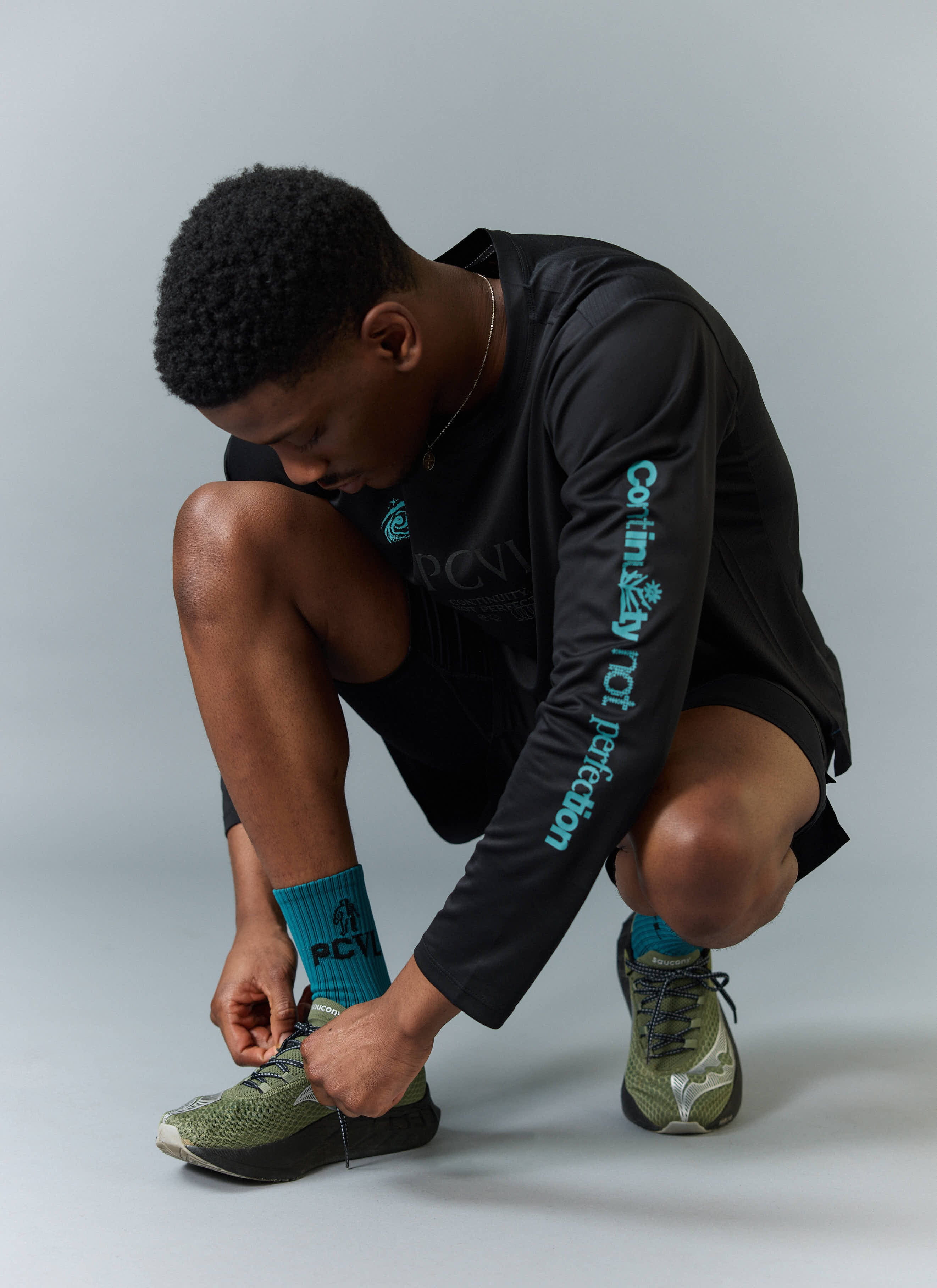 A model wearing the Percival black long sleeve PCVL performance top, crouching to tie the laces on his green trainers.