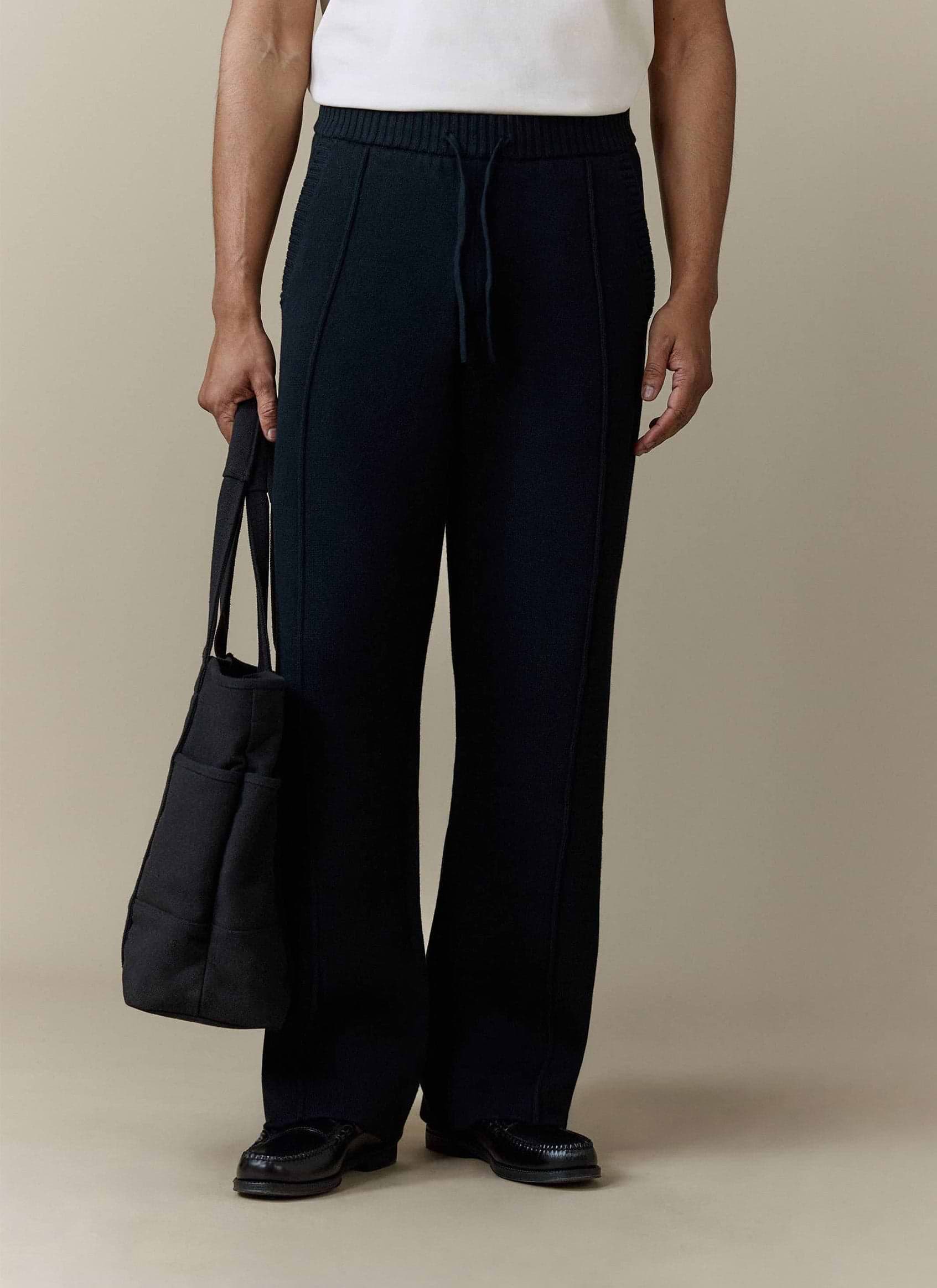 A model wears the Percival Chronicle Knitted Straight Leg Trackpant in black, styled with loafers and holding a tote bag.