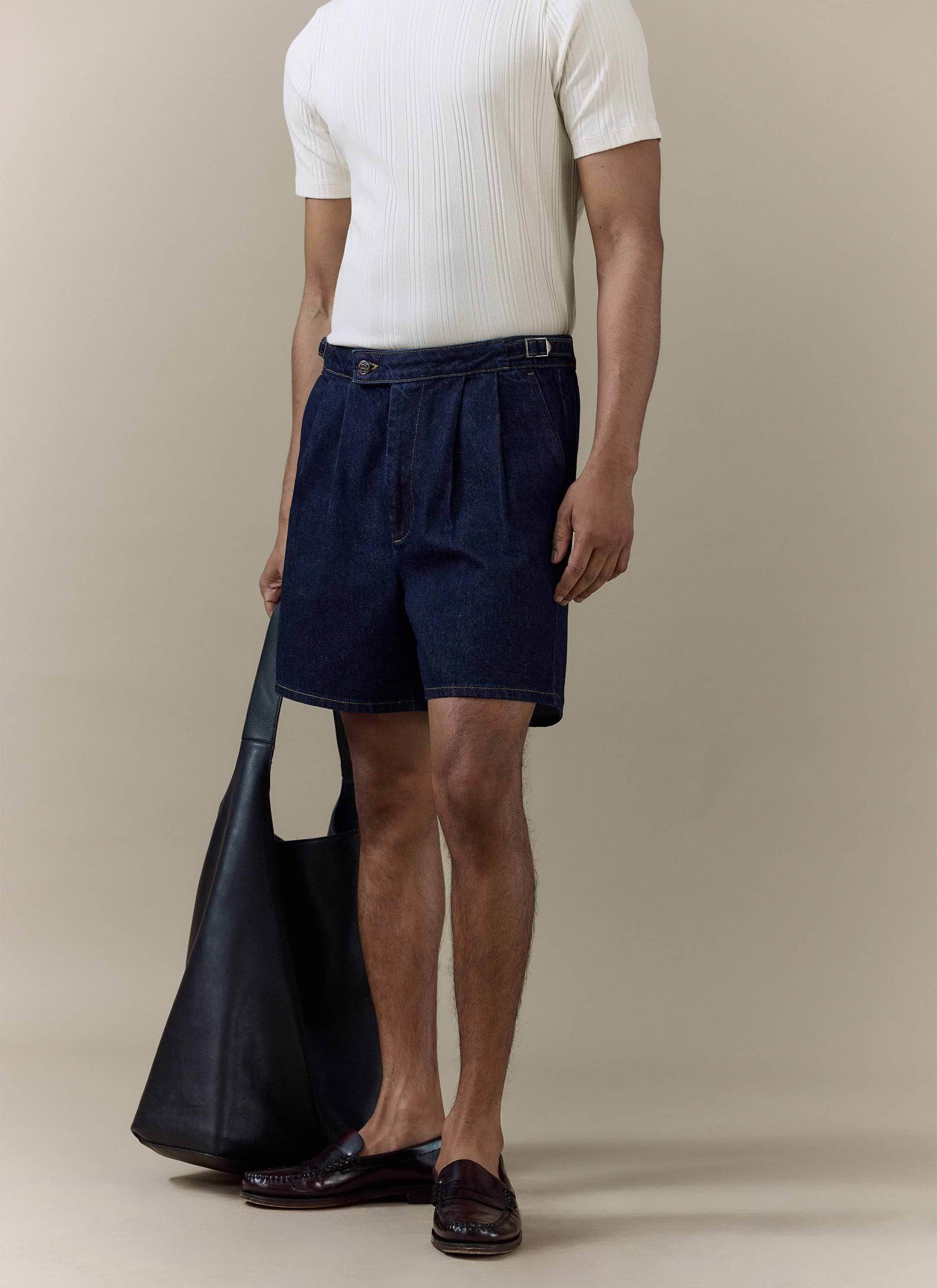 Percival Blake Double Pleated Denim Shorts in dark indigo cotton, styled on a model with a white tee and black leather bag.
