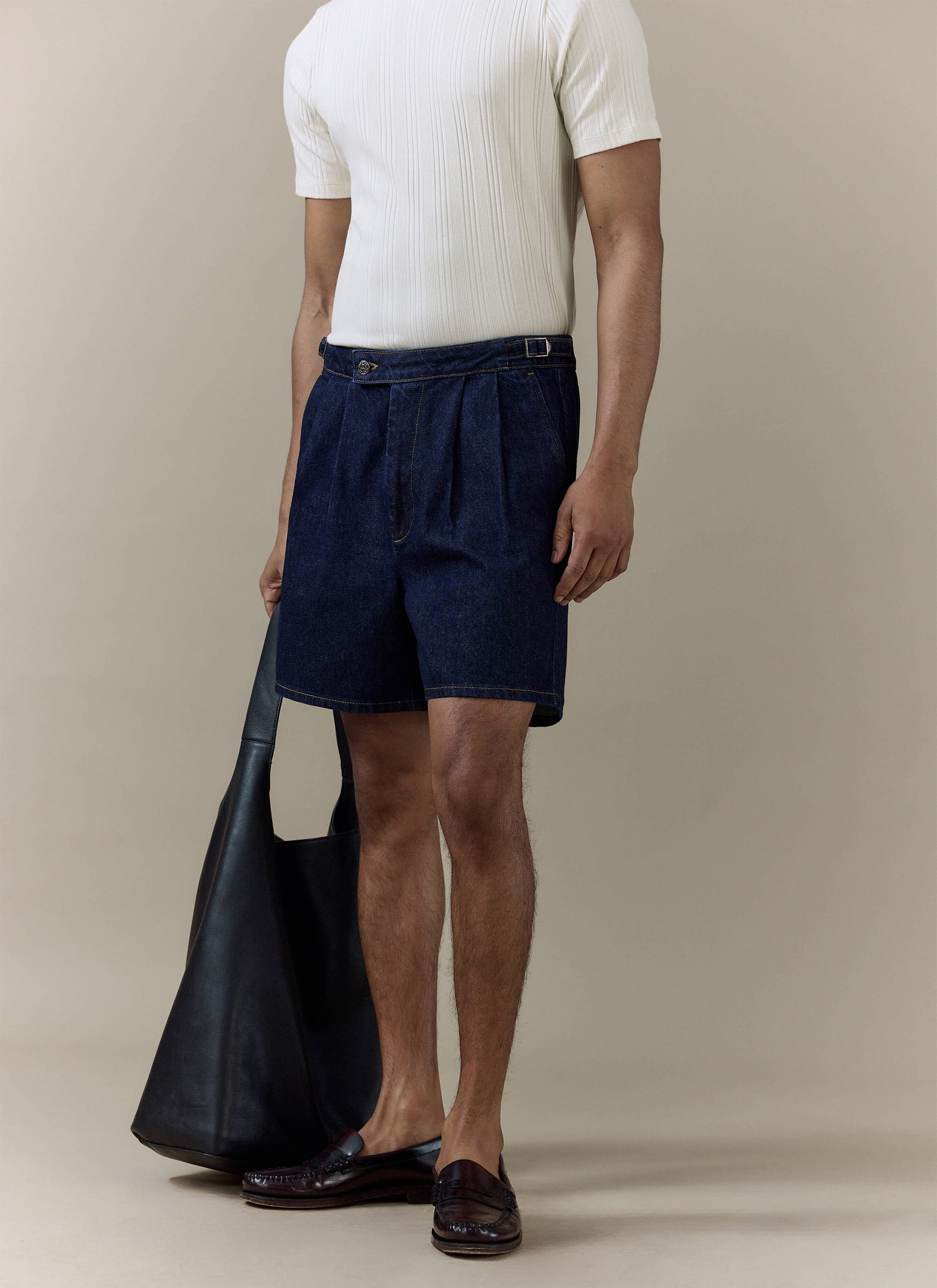 Percival Blake Double Pleated Denim Shorts in dark indigo cotton, styled on a model with a white tee and black leather bag.