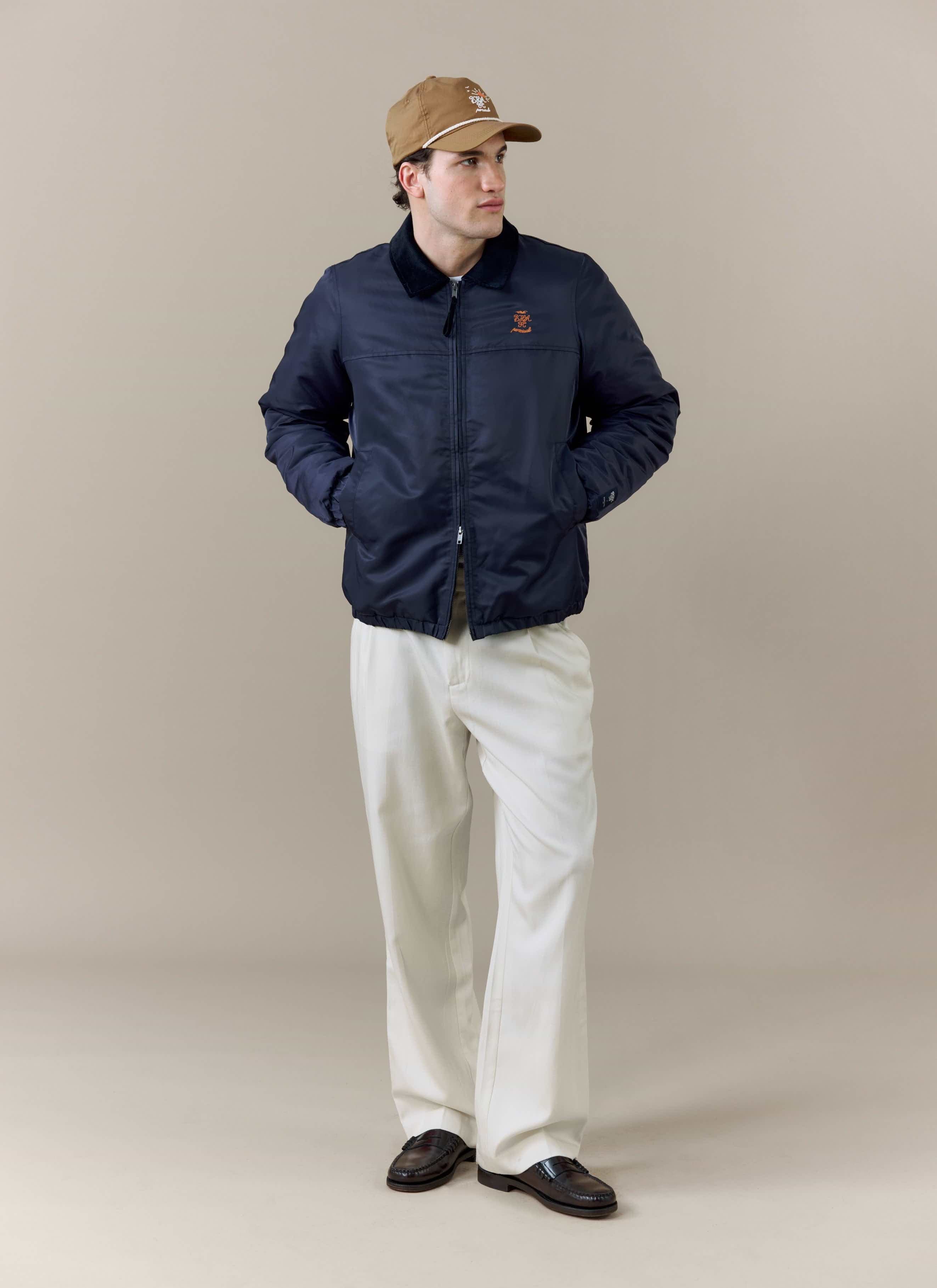 A model wears the Percival x Brighton & Hove Albion F.C. navy Coach Jacket, styled with white trousers and a tan cap.