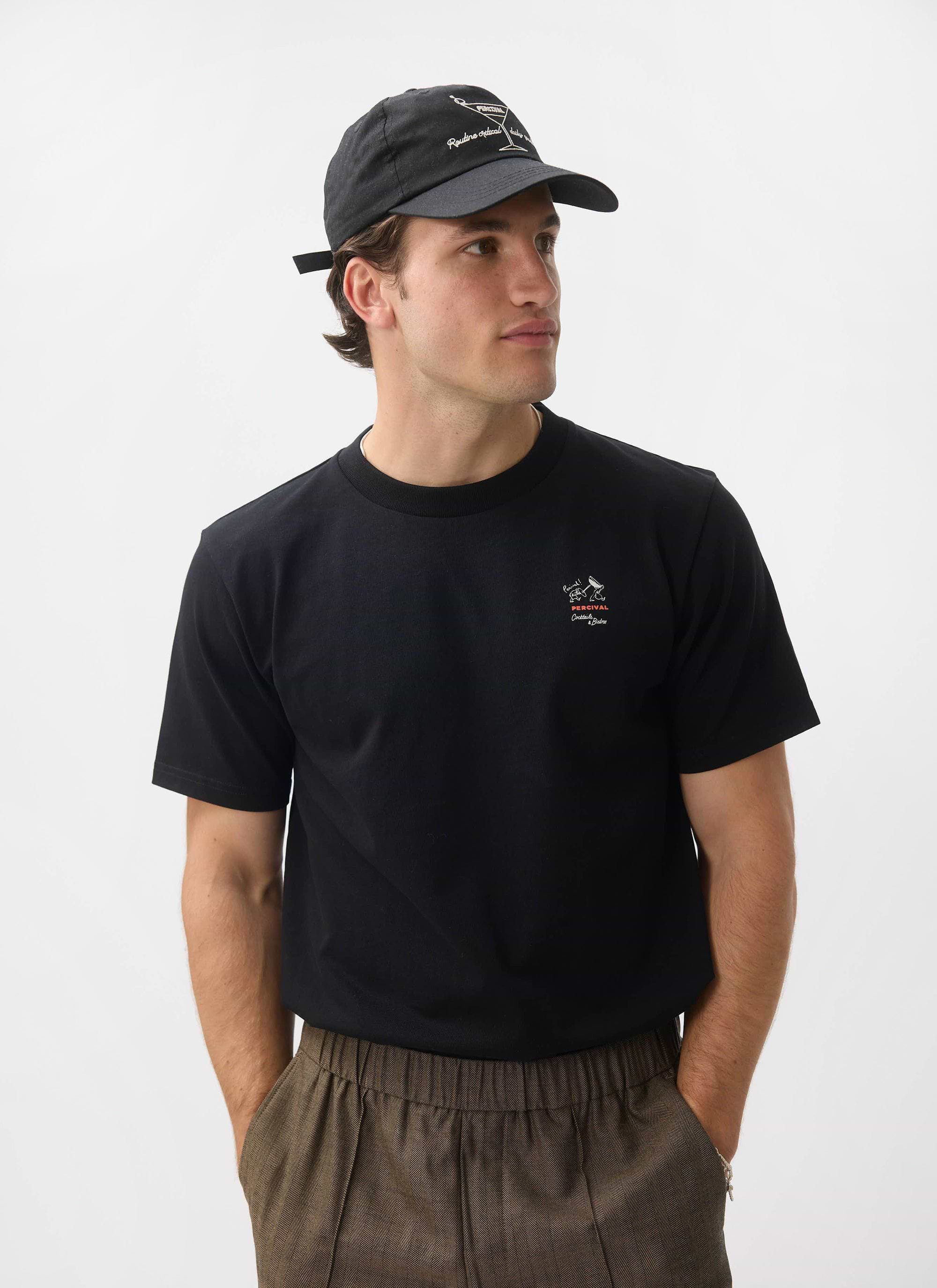 A model wears the black Percival À La Carte T-shirt, styled with brown trousers and a cap against a white studio background.
