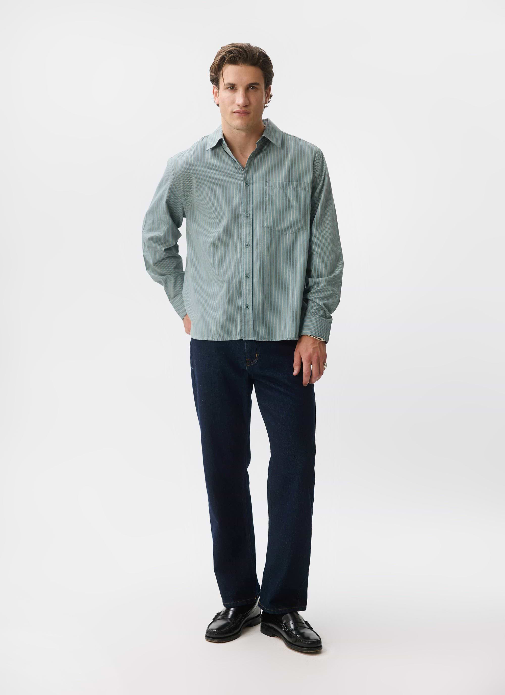 A man models the blue Percival Clancy Striped Long Sleeve Shirt, styled with dark denim and black loafers.