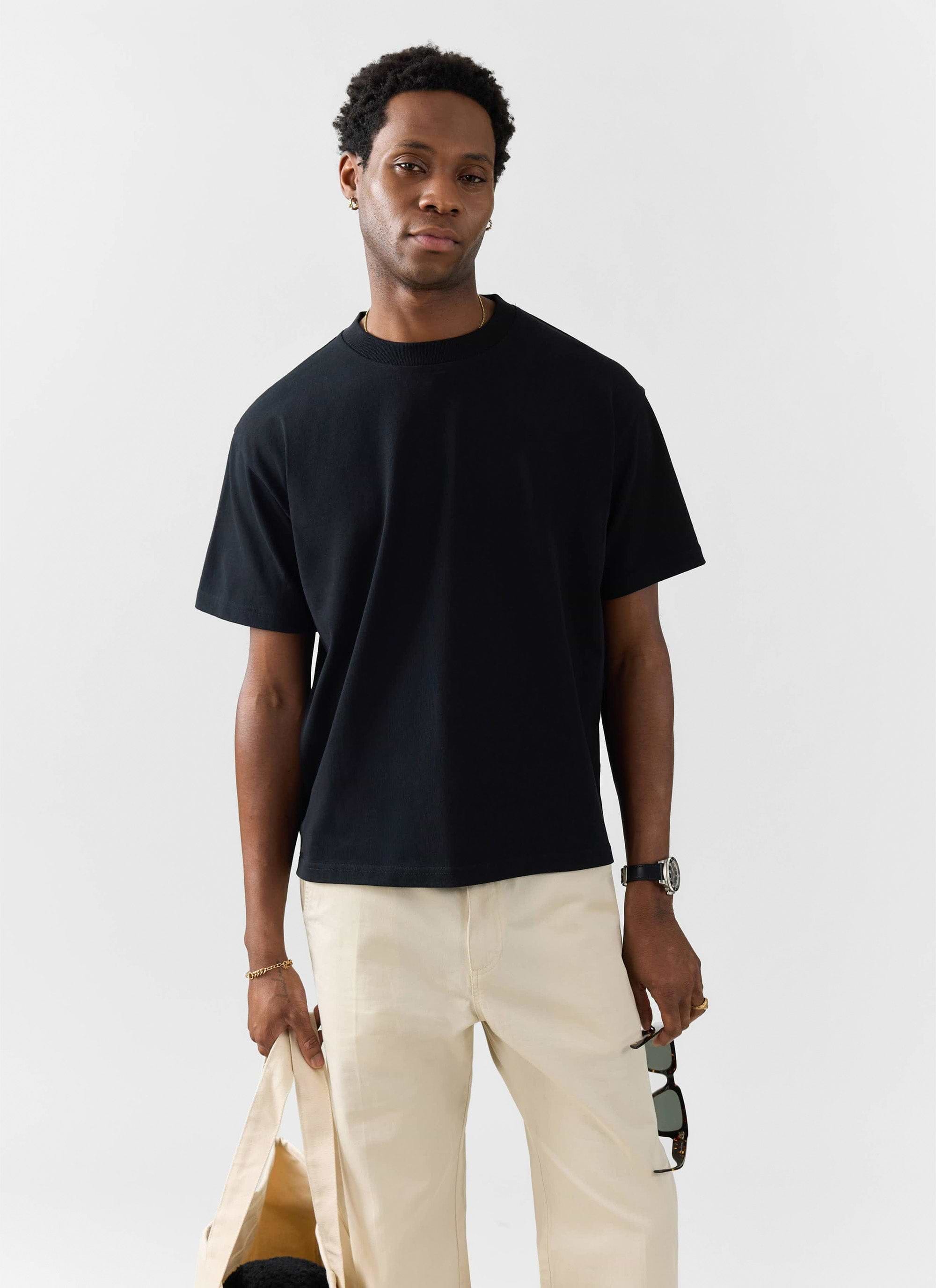 A man models the Percival black oversized t-shirt in organic cotton, styled with cream trousers against a white background.