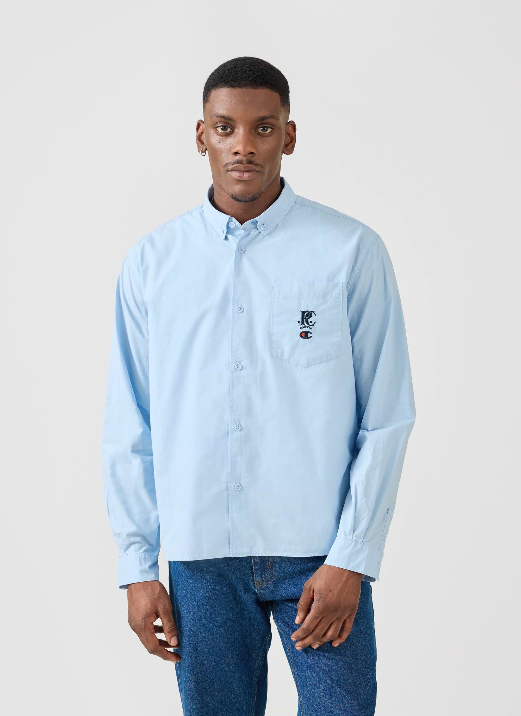 A model wears the light blue Champion x Percival Relaxed Oxford Shirt, a button-down style available from Percival.