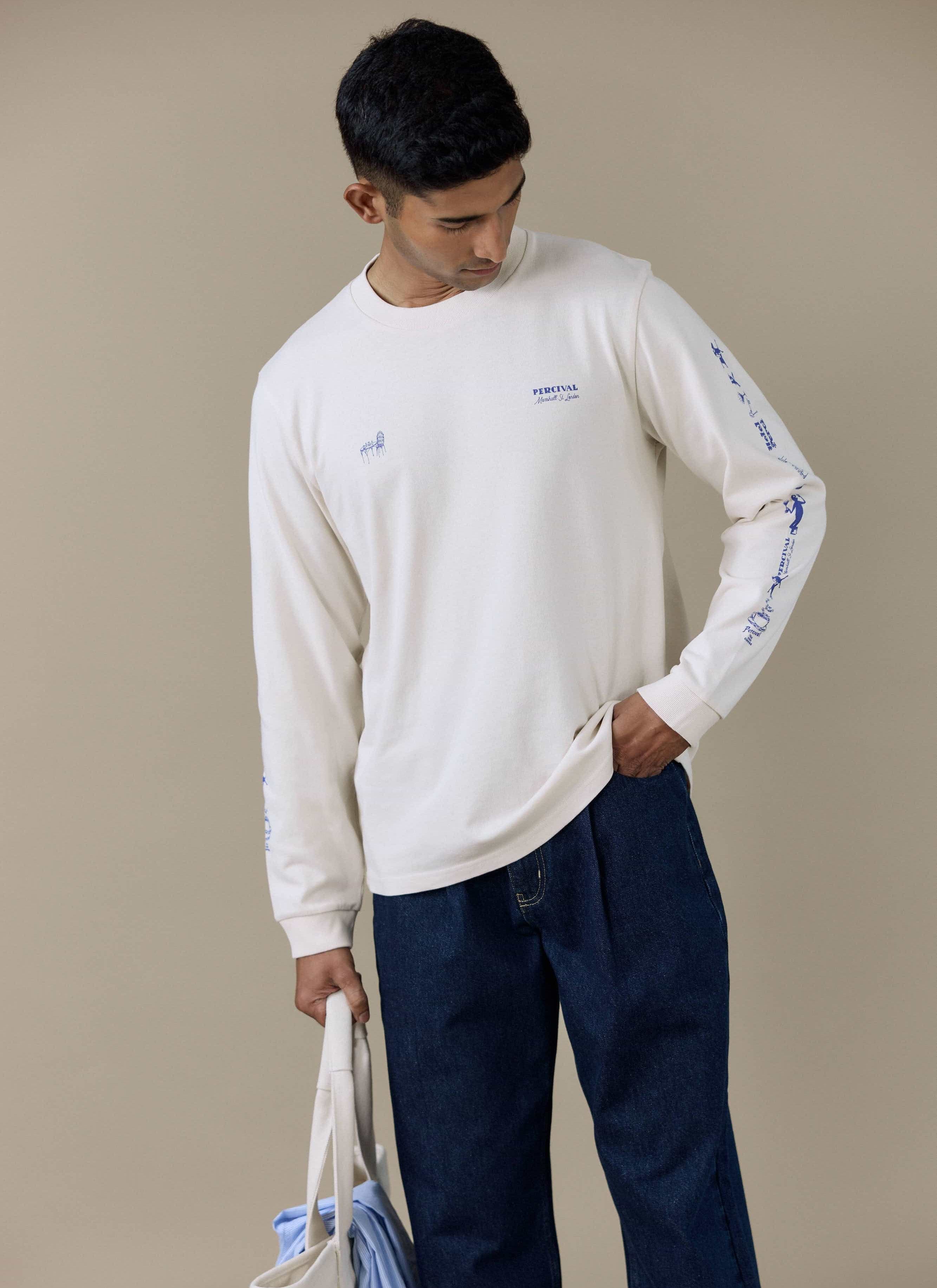 Model wearing the Percival Harlow Fine Wines Long Sleeve T Shirt in cream with blue graphics on the chest and sleeves.