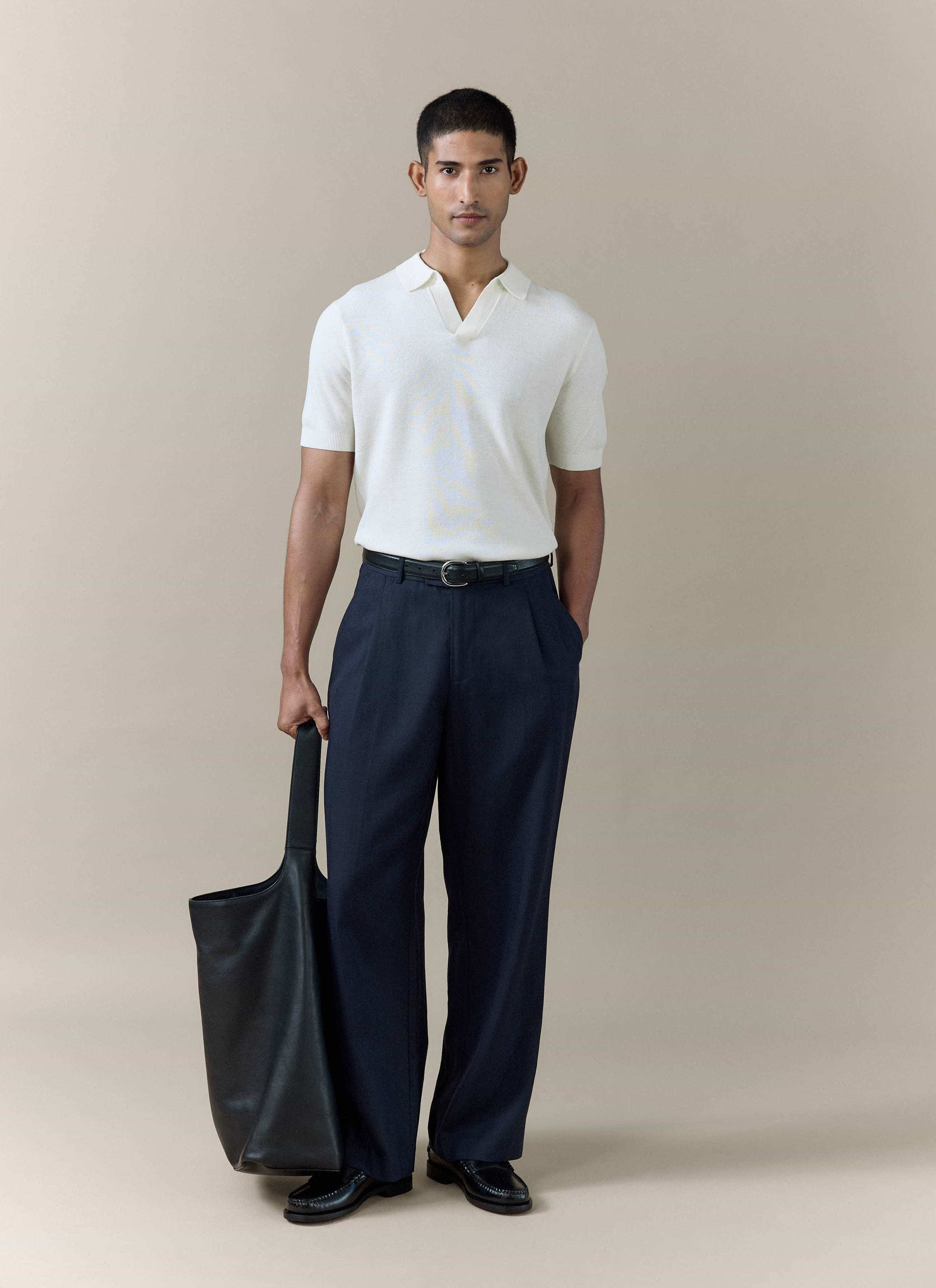 Percival Laurence cream knitted polo in a linen-lyocell blend, styled with navy pleated trousers and a black tote bag.