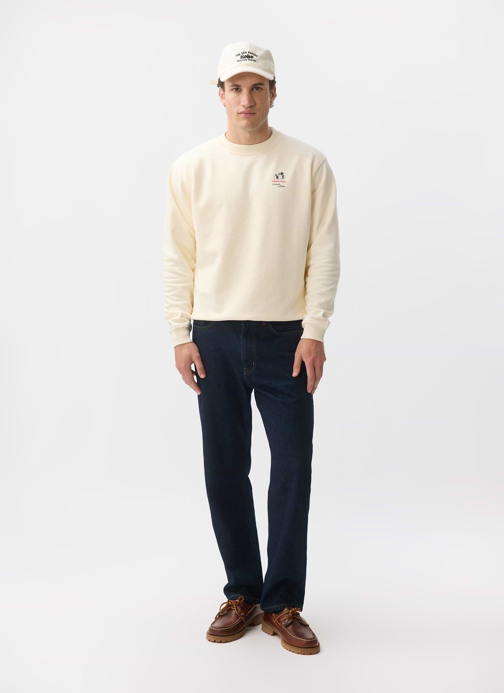 A model wears the Percival Bistro Printed Sweatshirt in ecru, styled with dark blue jeans, brown shoes and a matching cap.