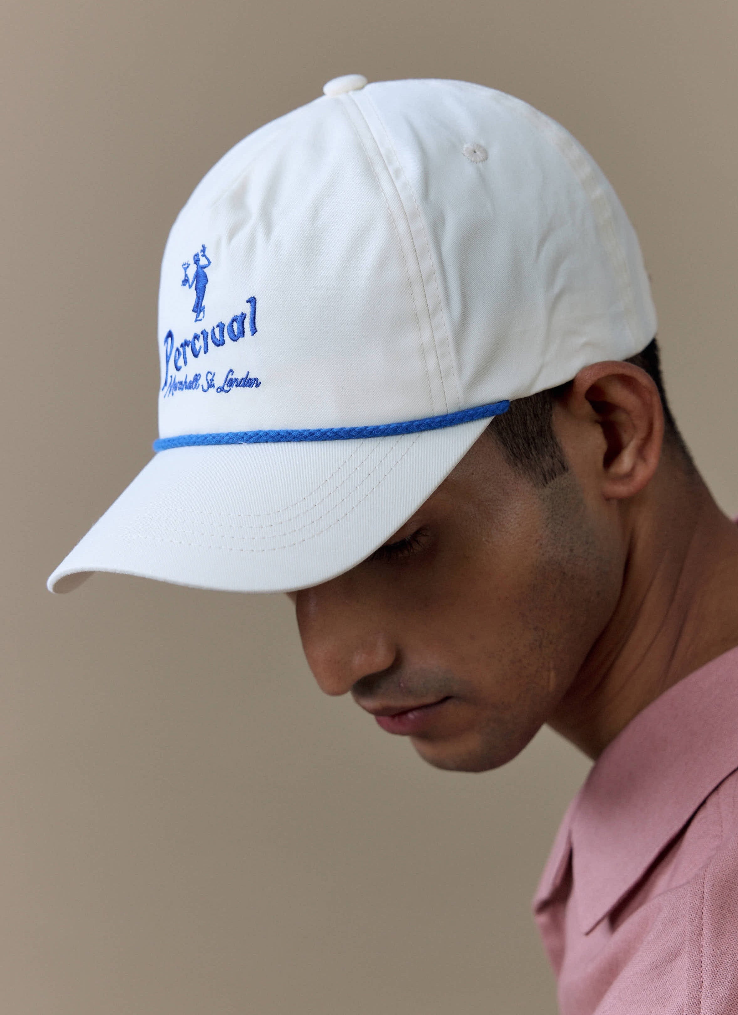 Close-up of a model wearing the Percival Martini Soft Trucker Cap in cream, featuring blue embroidery and rope detail.