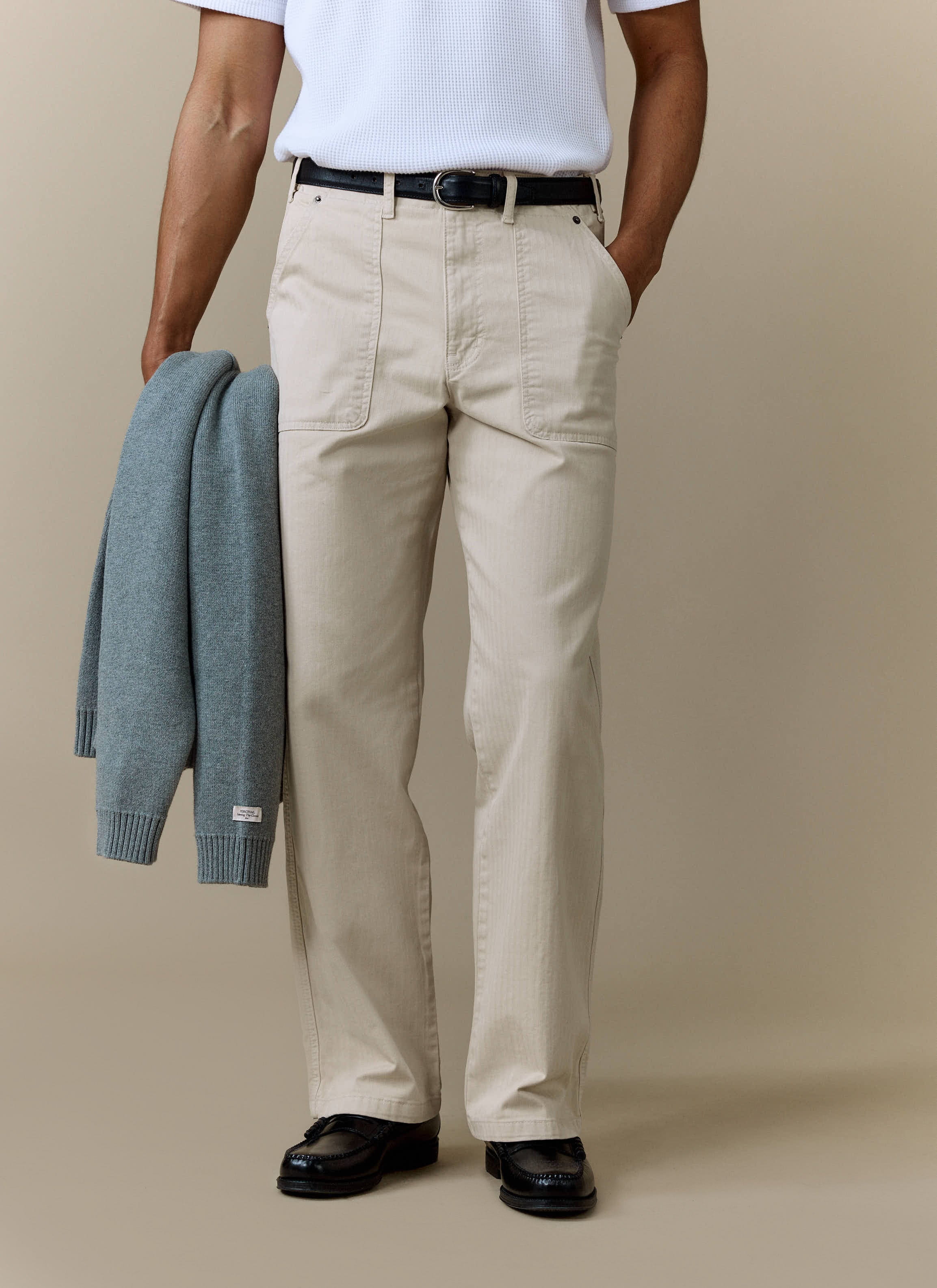 A model wearing Percival Lungo Herringbone Straight Trousers in cream, styled with a white tee and blue cardigan.