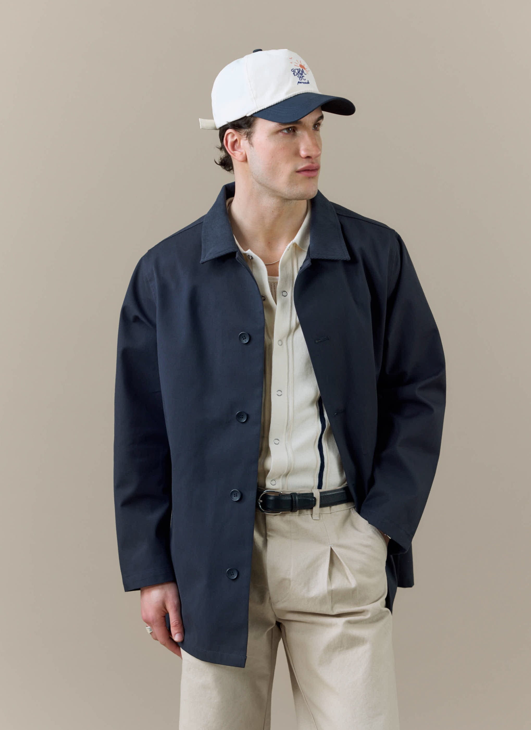 A model wears the Percival x Brighton & Hove Albion F.C. Sherlock Trench in Midnight, styled with a cap and cream trousers.