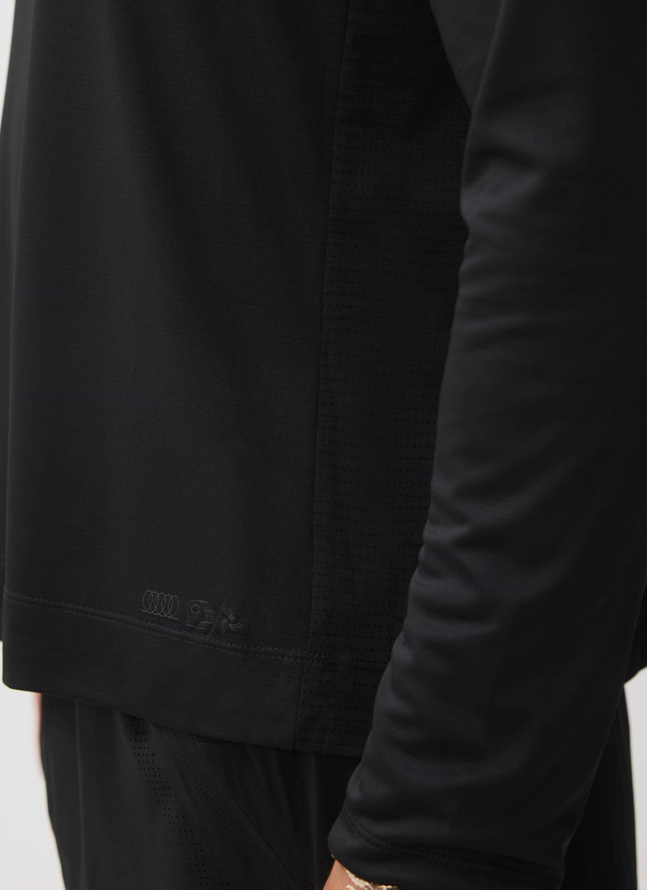 A close-up of the Percival PCVL Long Sleeve Performance Top in black, showing the fabric detail and tonal logo at the hem.