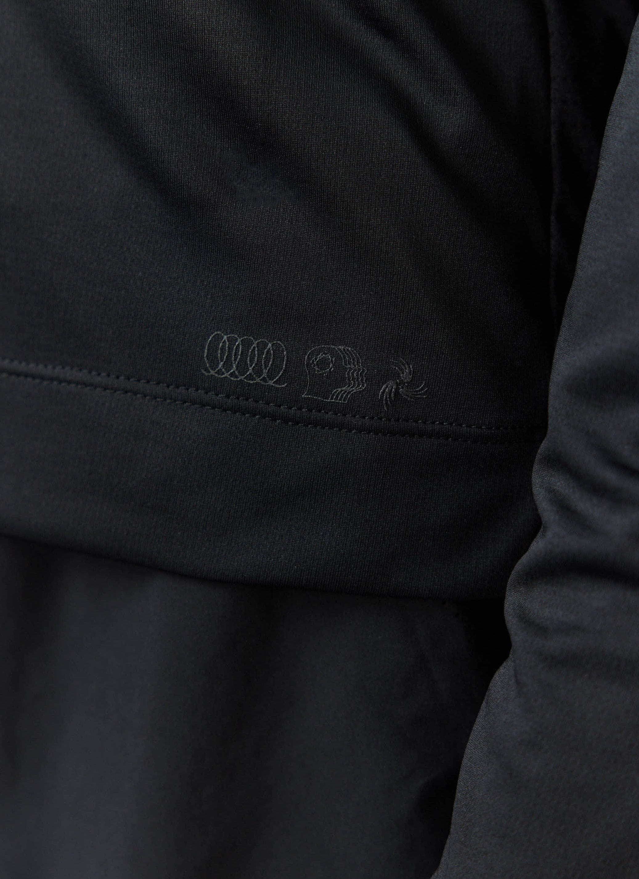 A close-up of the tonal PCVL graphic embroidered on the black Long Sleeve Performance Top by Percival.