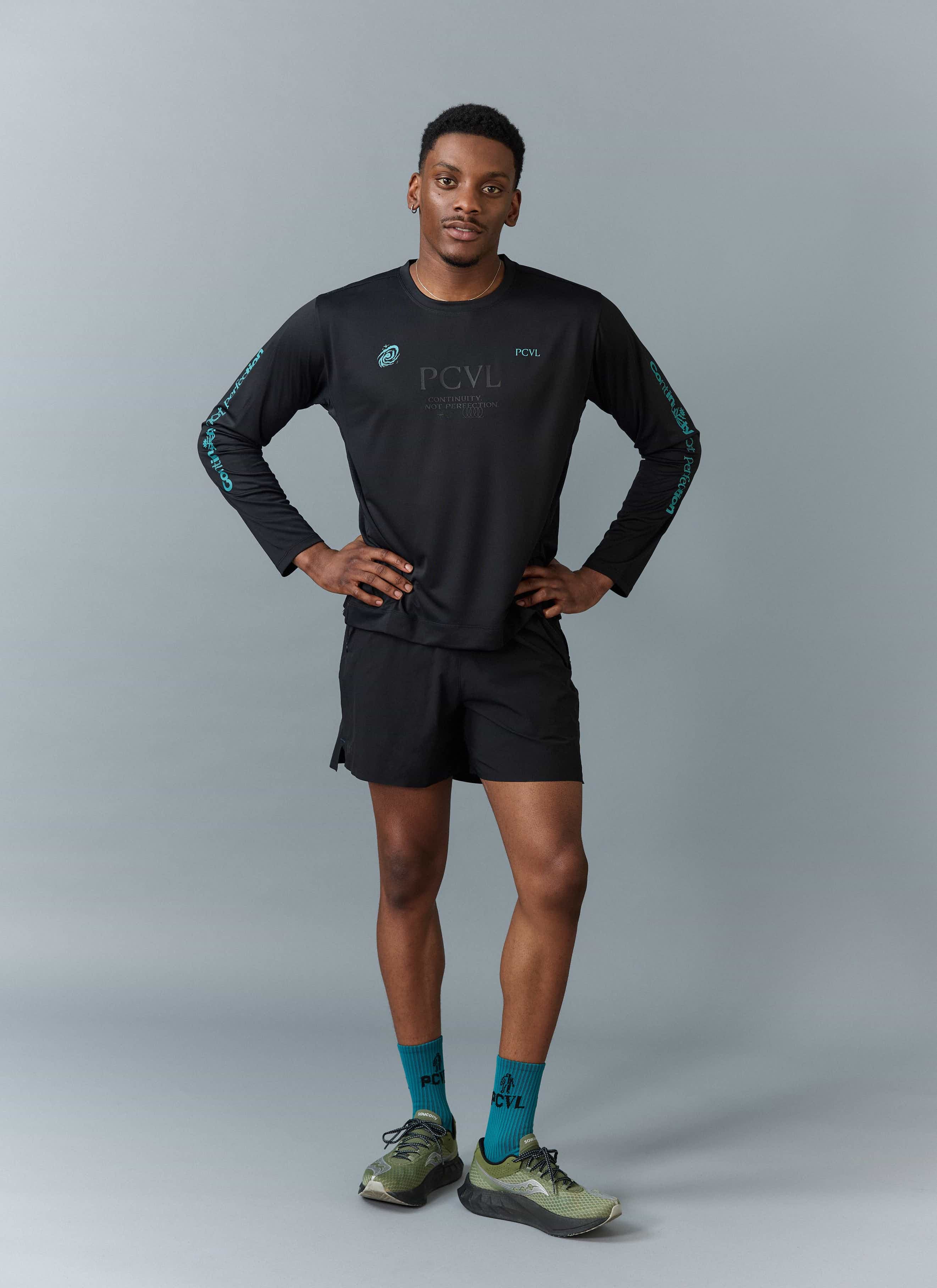 A model wears the black PCVL Long Sleeve Performance Top by Percival with matching shorts against a grey studio background.