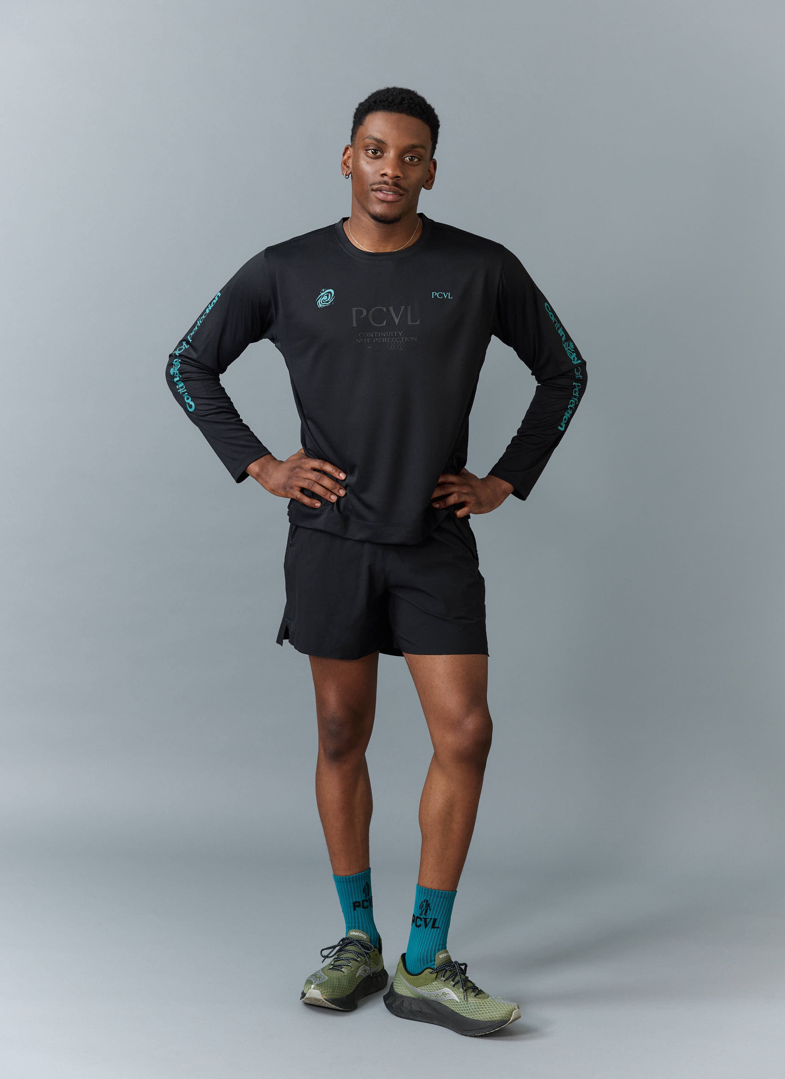 A model wears the black PCVL Long Sleeve Performance Top by Percival with matching shorts against a grey studio background.