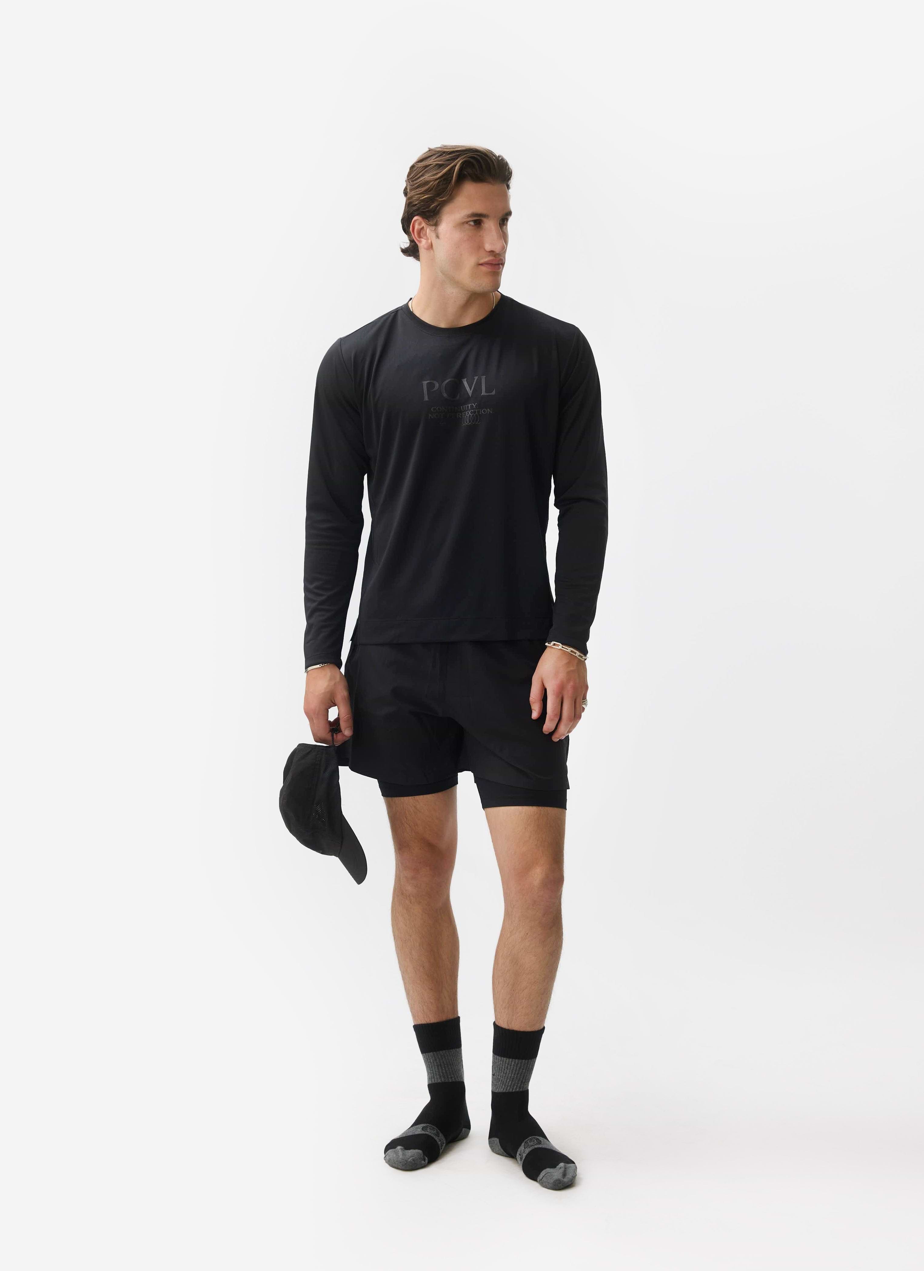 A model poses in a studio wearing the Percival PCVL Long Sleeve Performance Top in black with matching shorts.