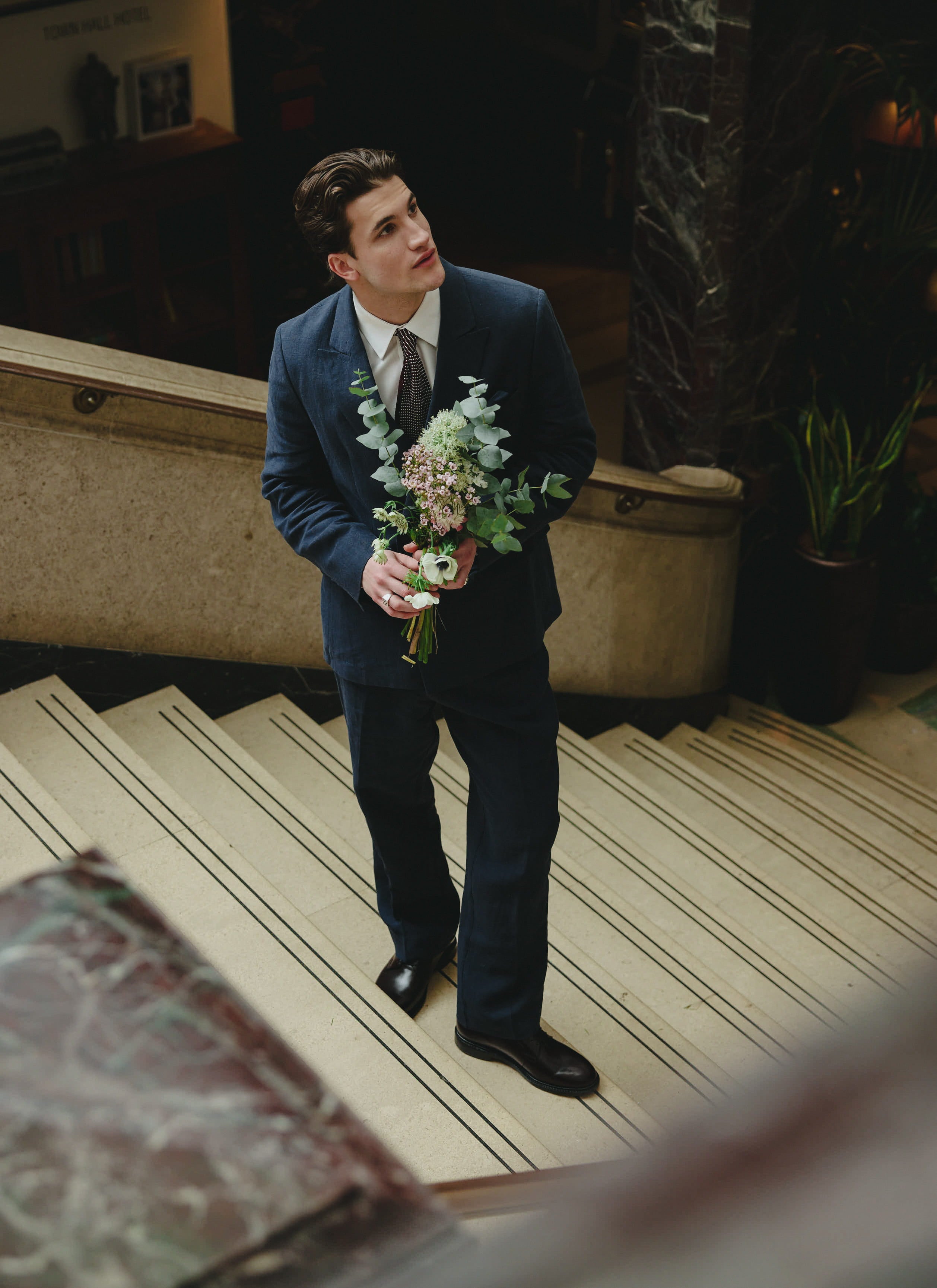 Percival Clifton double breasted blazer in navy blue linen, worn with trousers and a bouquet on a grand staircase.