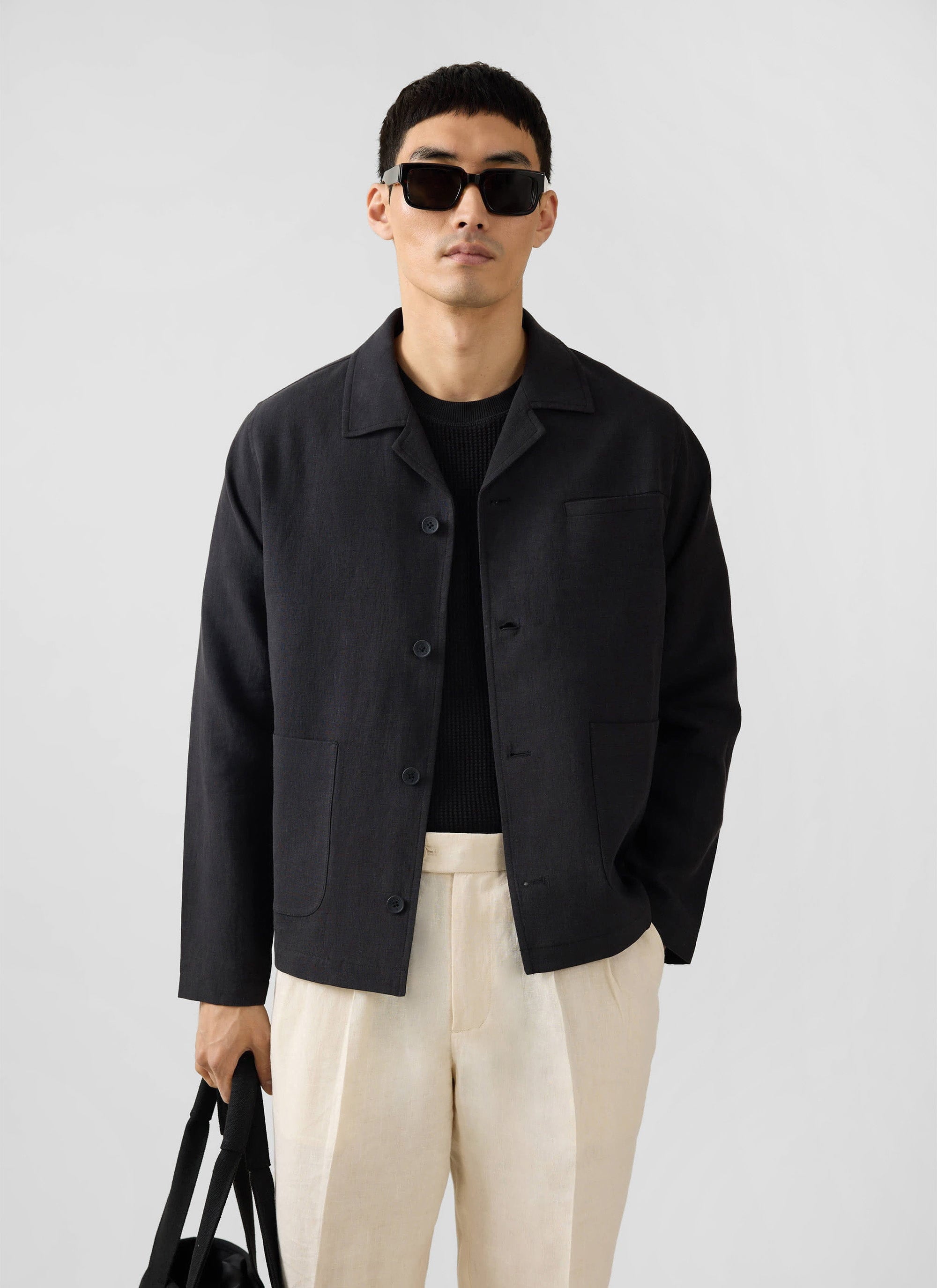 A model wears the Percival black linen overshirt, styled with cream trousers and sunglasses in a studio.