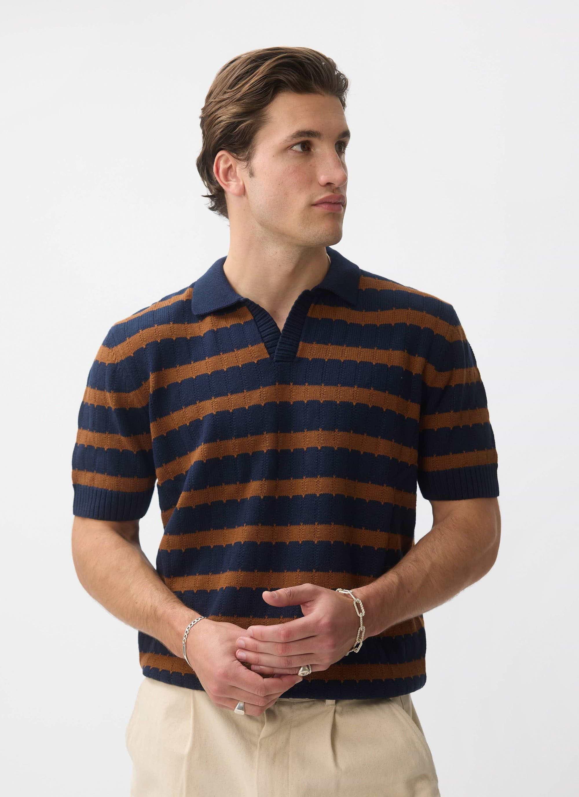 A male model wearing the Percival Stine Knitted Polo in a brown and navy stripe, standing in a studio.