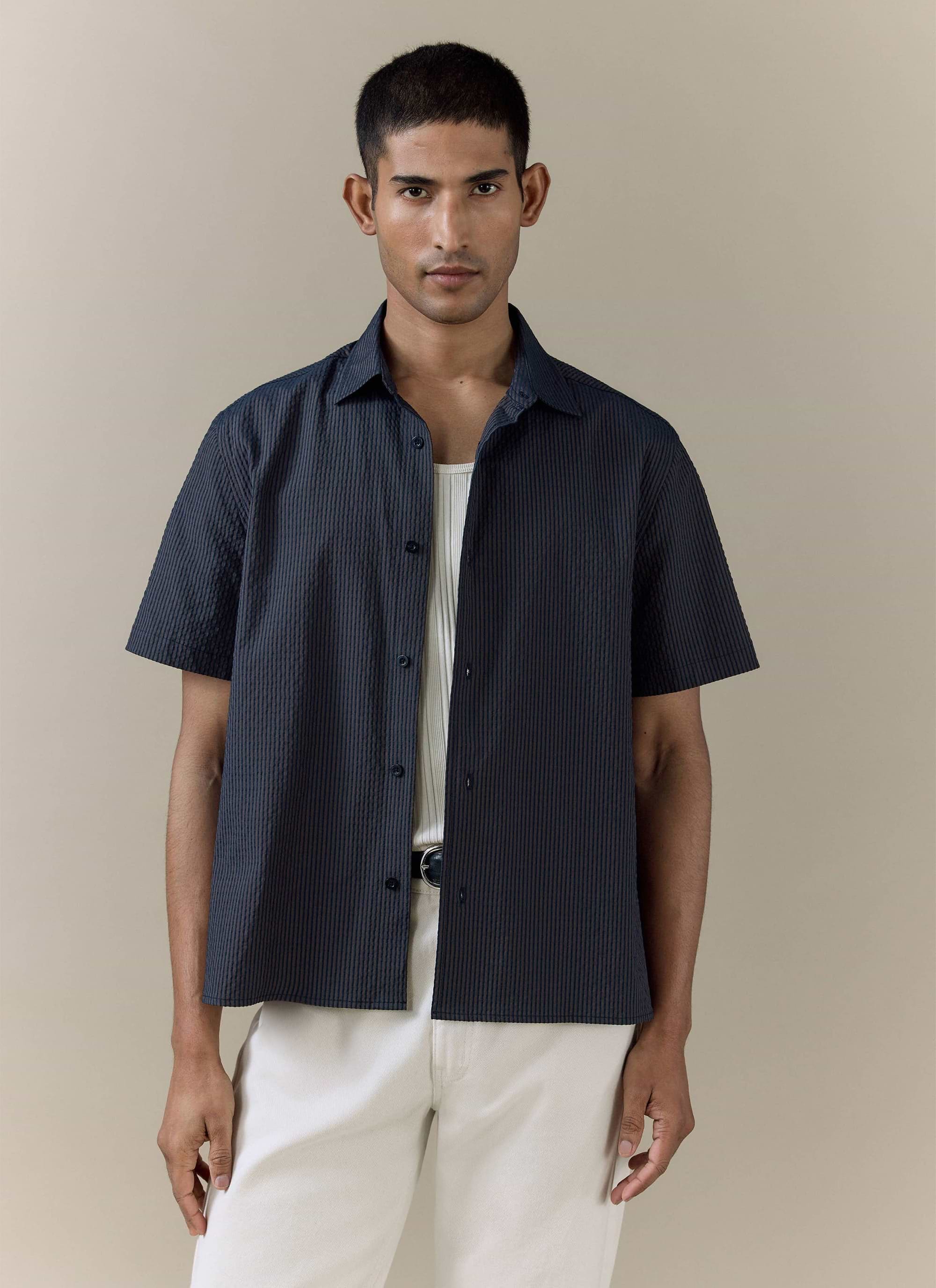 Percival Rowan Seersucker Boxy Shirt in navy stripe cotton, styled open over a white ribbed vest and cream trousers.