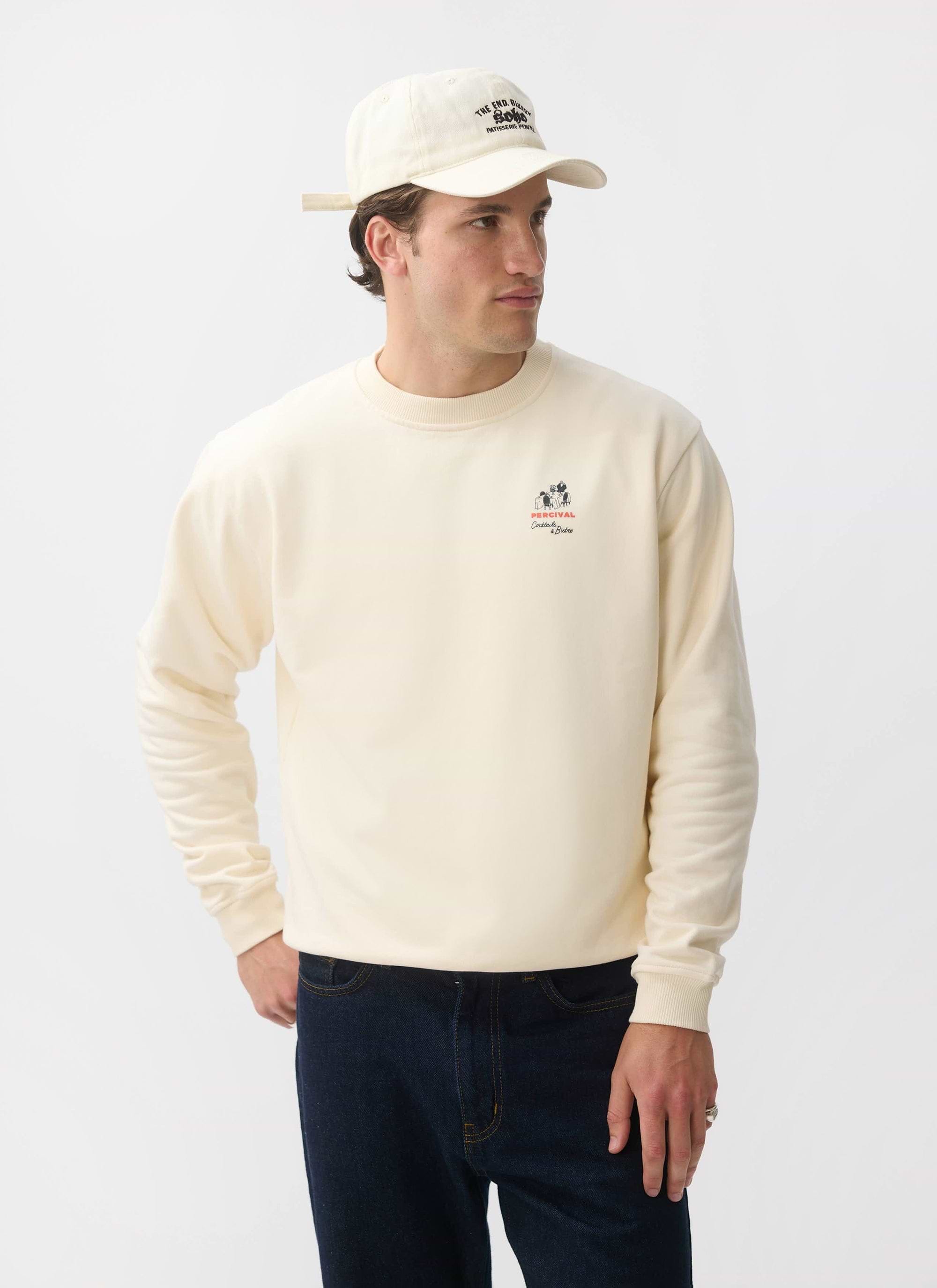 A model wears the Percival Bistro Printed Sweatshirt in ecru, styled with dark jeans and a baseball cap.
