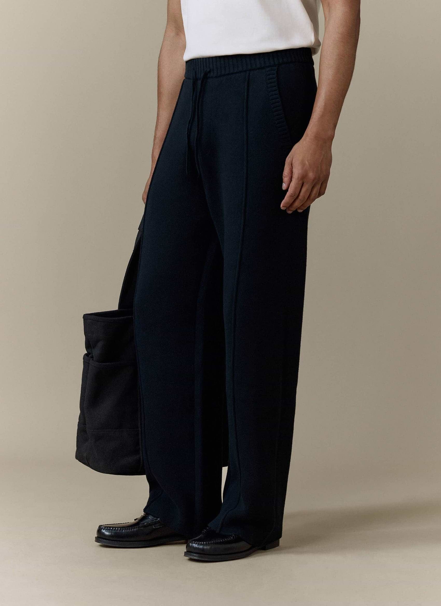 A model wears the black Chronicle Knitted Straight Leg Trackpants by Percival, styled with a tote bag and black loafers.