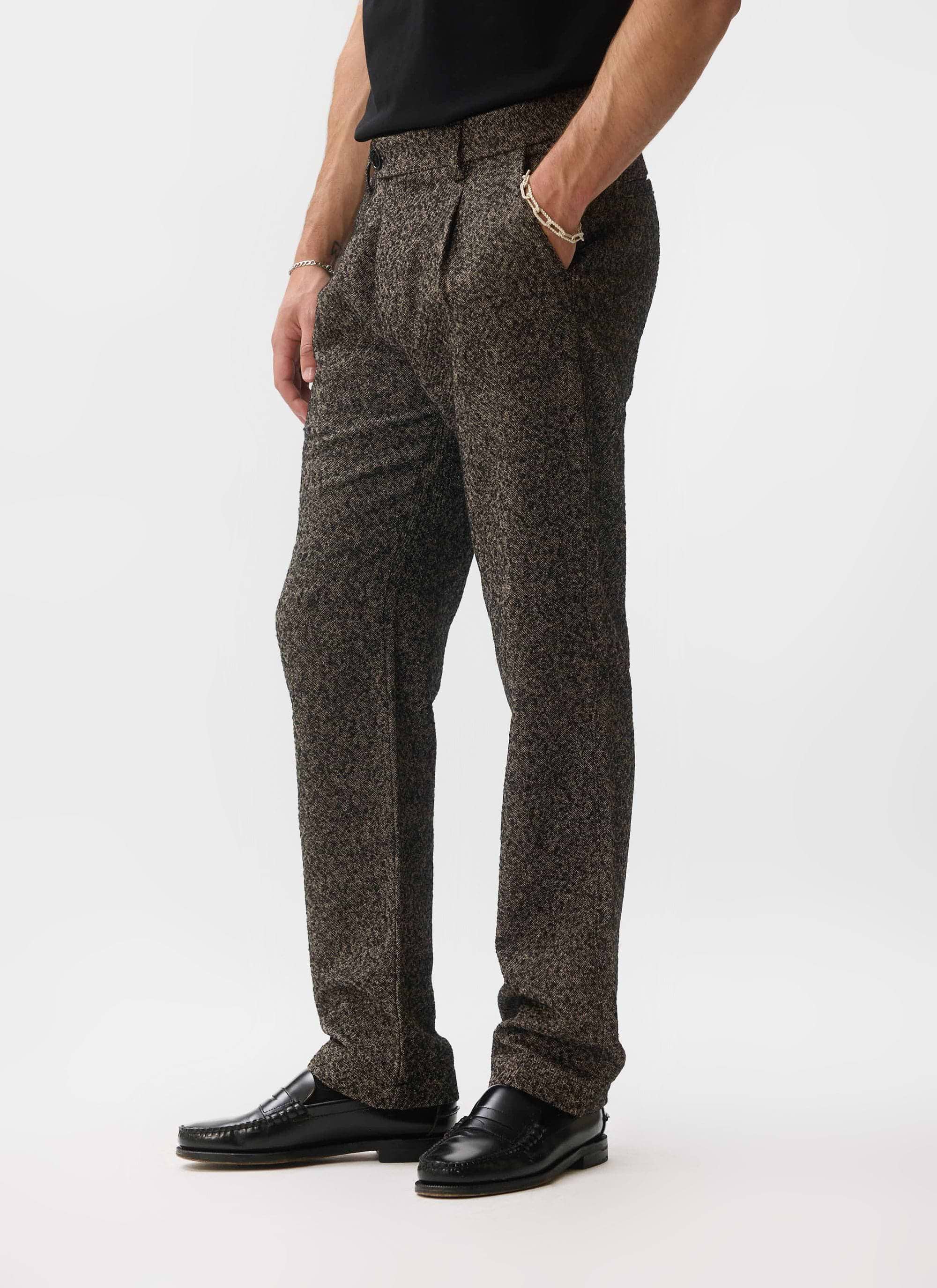 A model wears the Percival Blyton Trousers in a grey nep fabric, styled with a black t-shirt and black loafers.