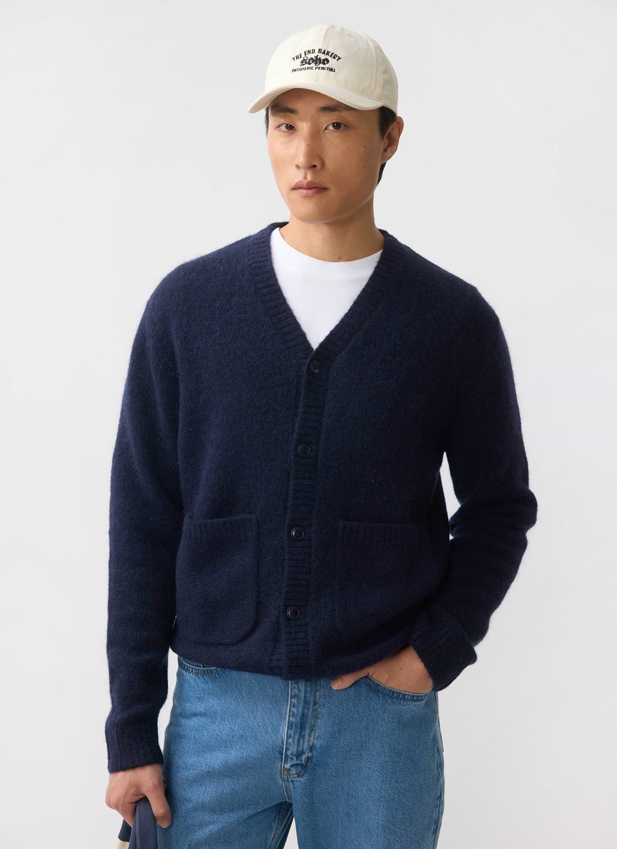 A man models the navy White Glove Cardigan from Percival, a soft mohair blend knit, styled with a white tee and baseball cap.
