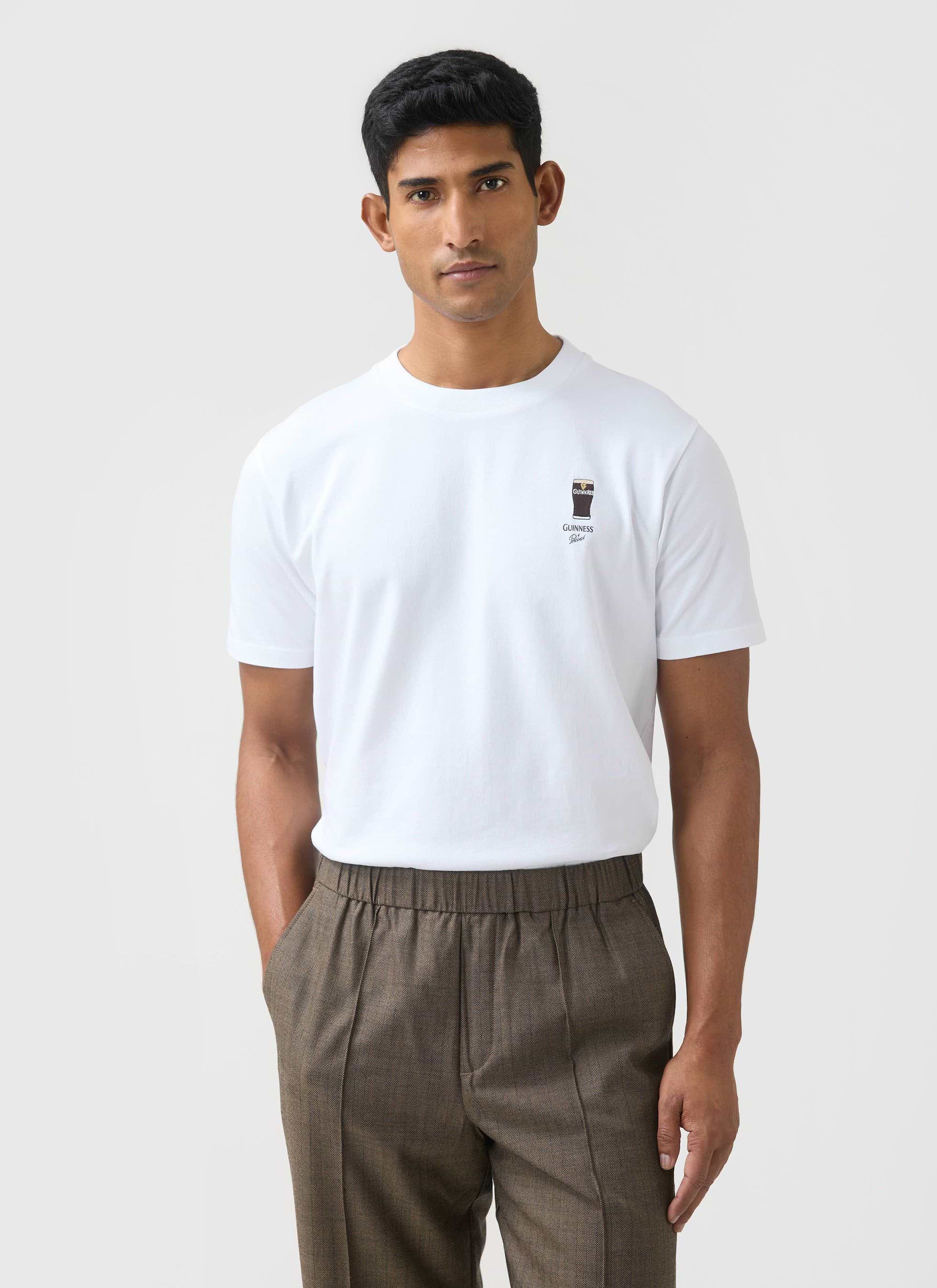A model wears the white Stout T-Shirt from the Percival x Guinness collection, styled with brown herringbone trousers.