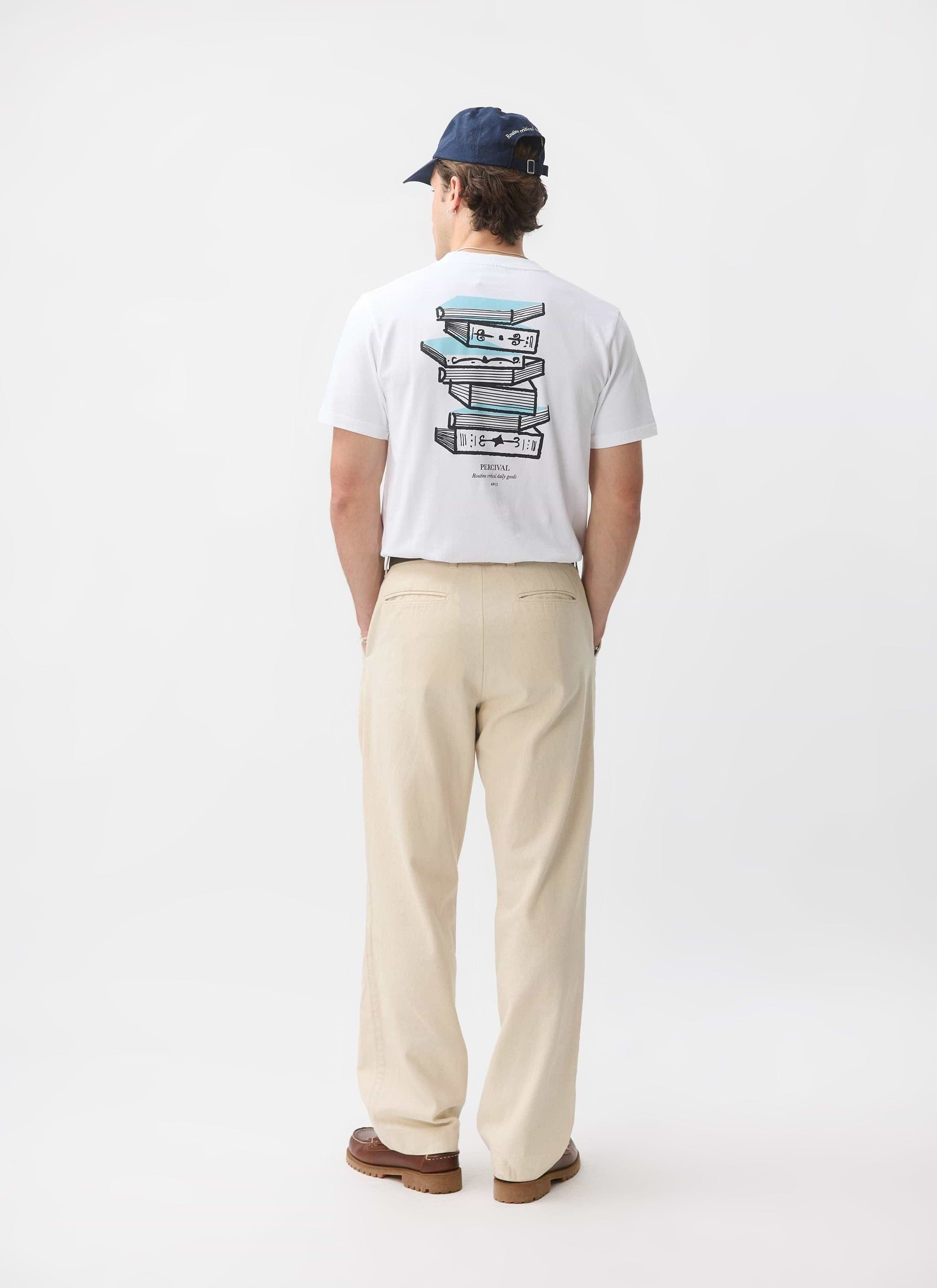 A model from behind wearing the Percival Duke T-shirt in white, showing the 'Routine Critical Daily Goods' book graphic.