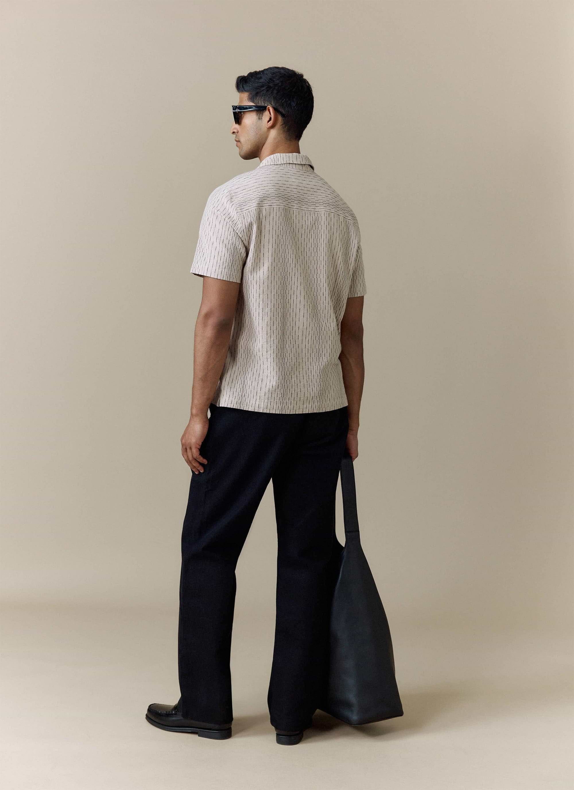 Rear view of the cream Percival Leland Cuban Shirt, styled with black trousers and a large leather tote bag.