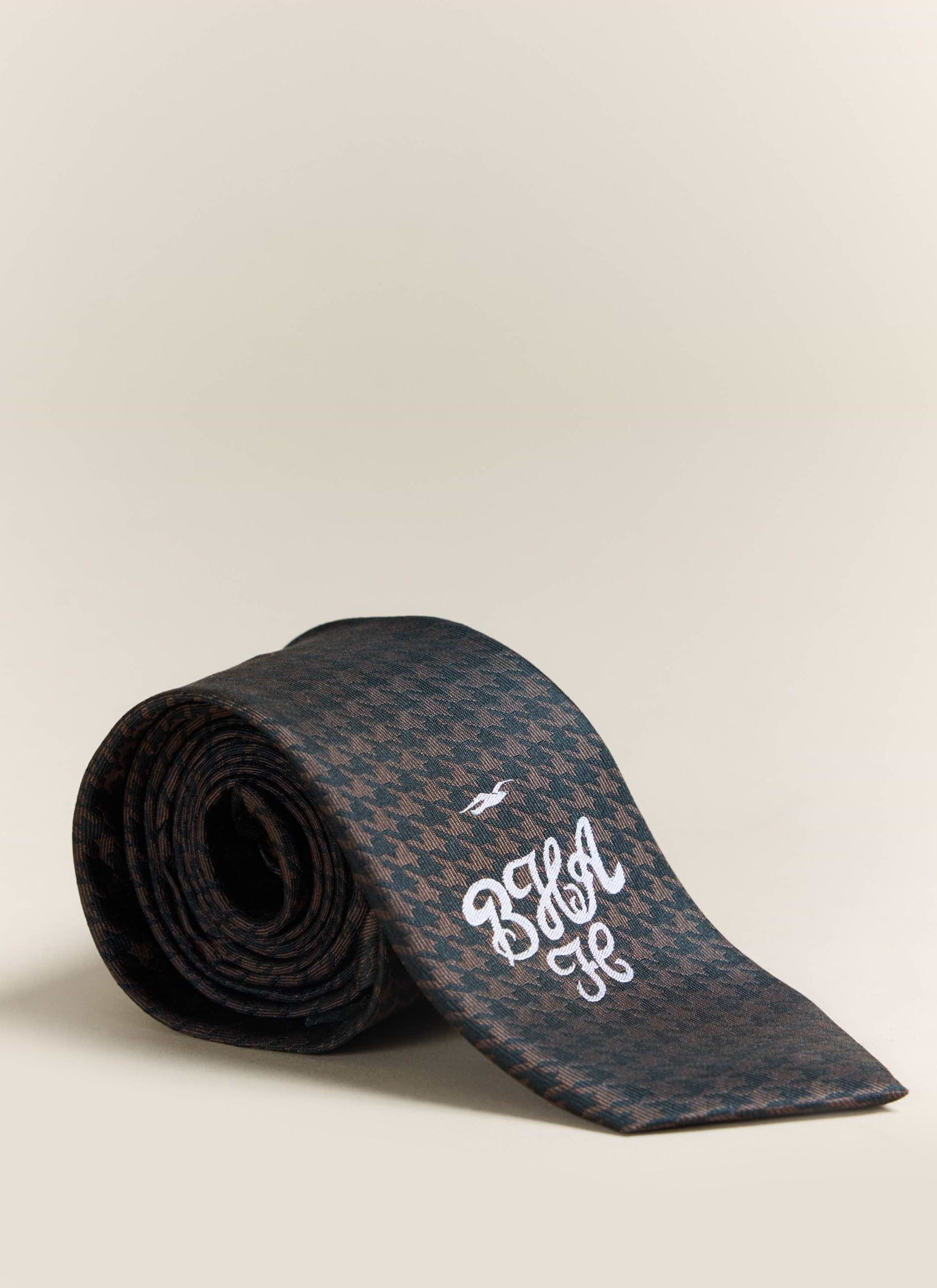 A rolled up Percival x Brighton & Hove Albion F.C. tie in brown silk, featuring a houndstooth pattern and embroidered logo.